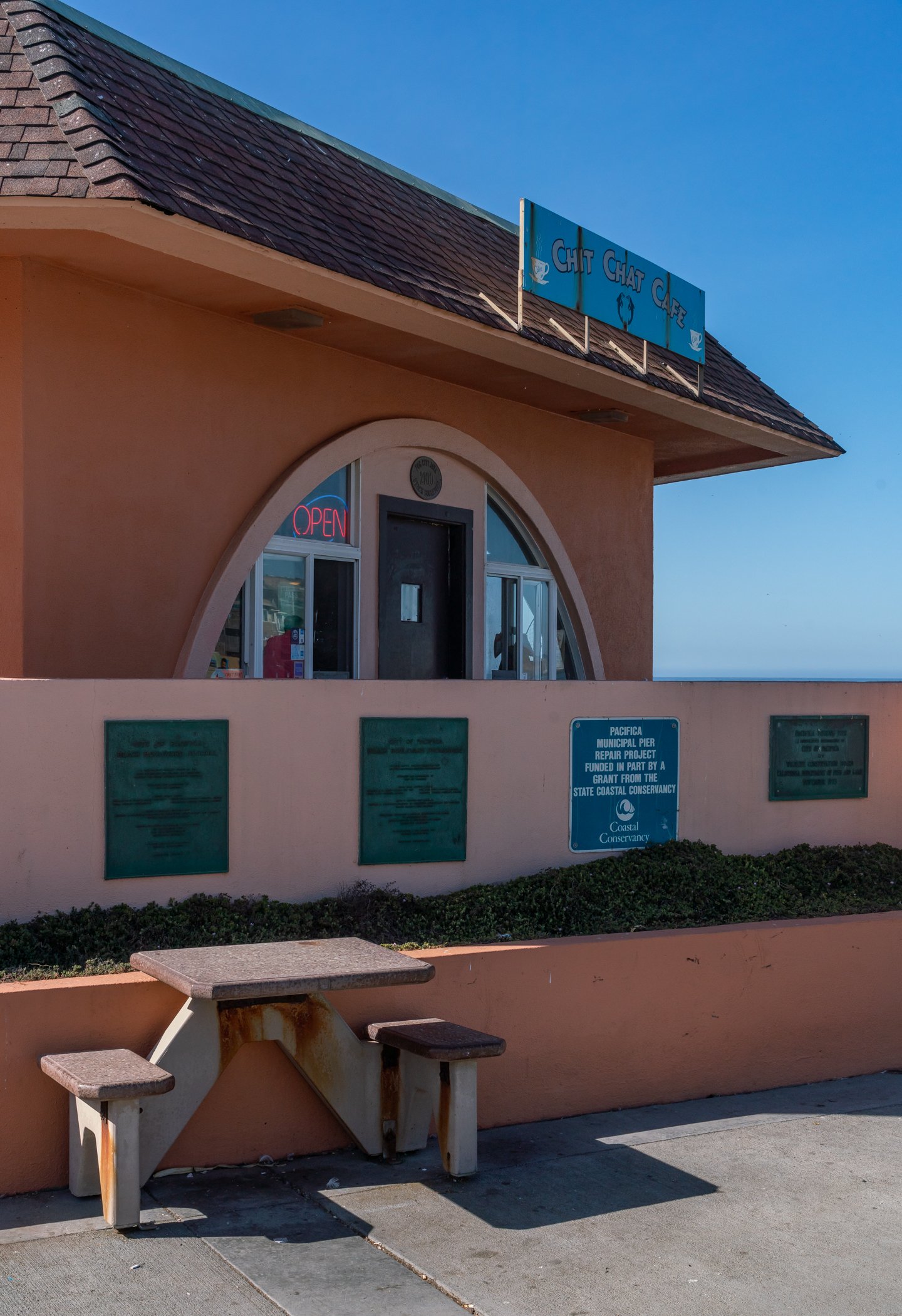 Building with a sign reading "Chit Chat Café" and "Open" sign in window, with a picnic table and informational plaques outside.