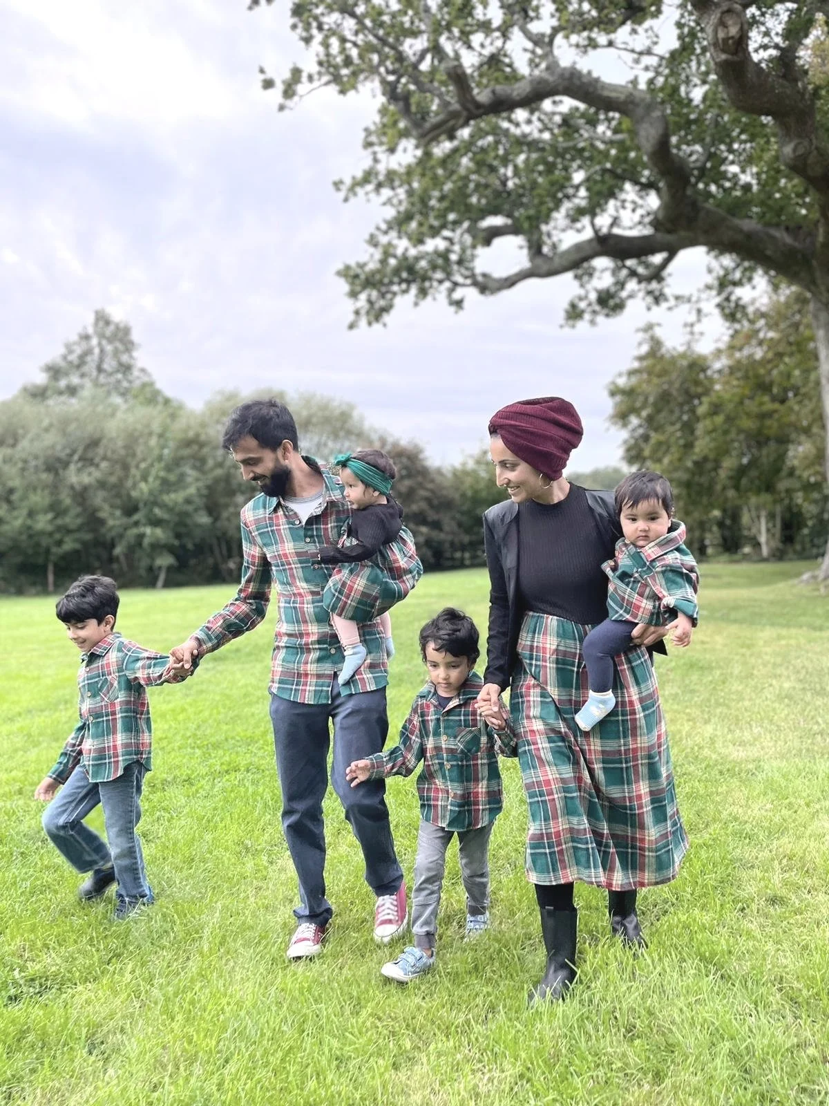 A family of five walking on grass, wearing matching plaid outfits. Two adults hold hands with three children. Trees and a cloudy sky are in the background.