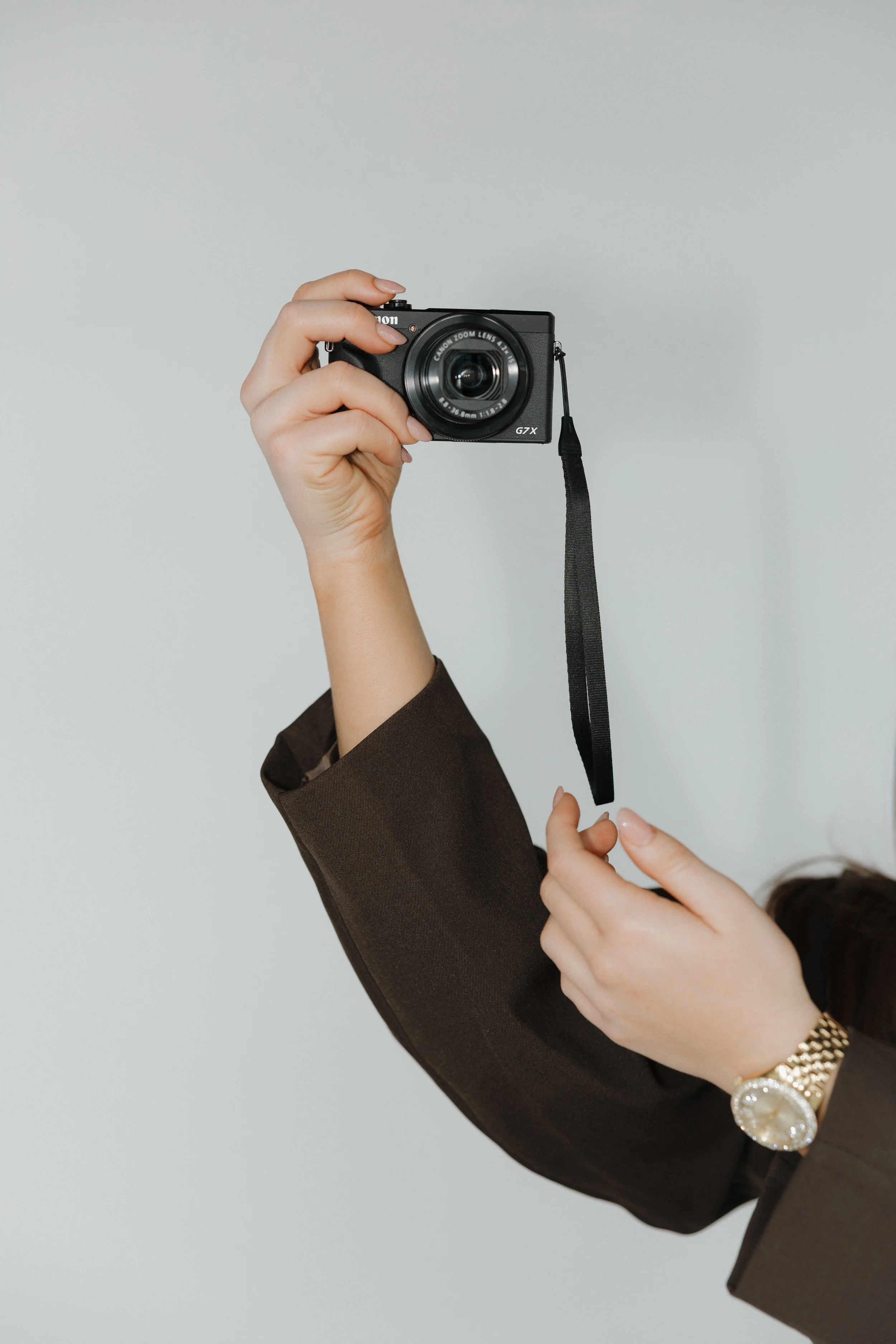 Person holding a small black digital camera with a strap, wearing a brown jacket and a gold watch. content creation for brands at the social current