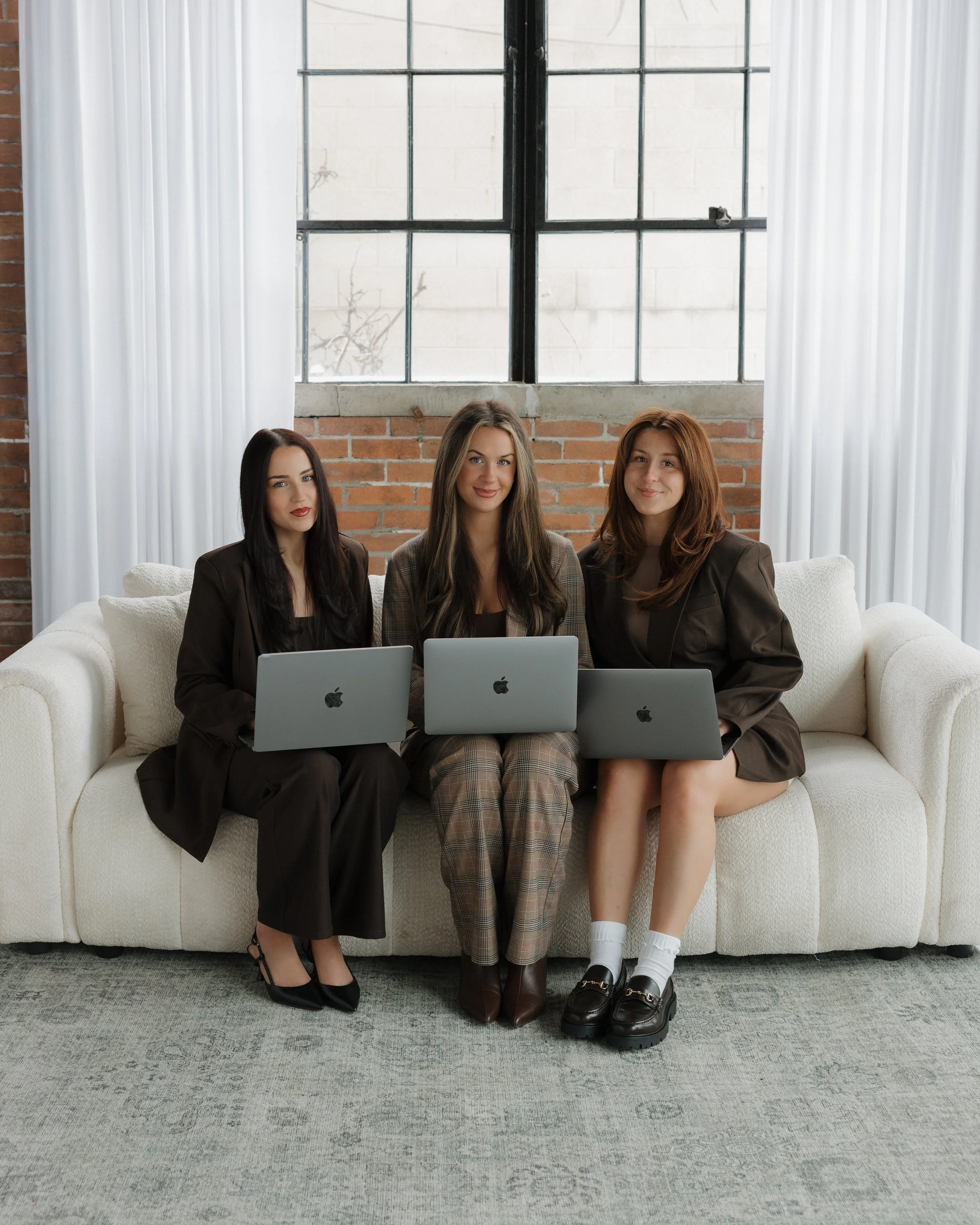 Three women sitting on a white sofa with laptops, in an industrial-style room with brick walls and large window.