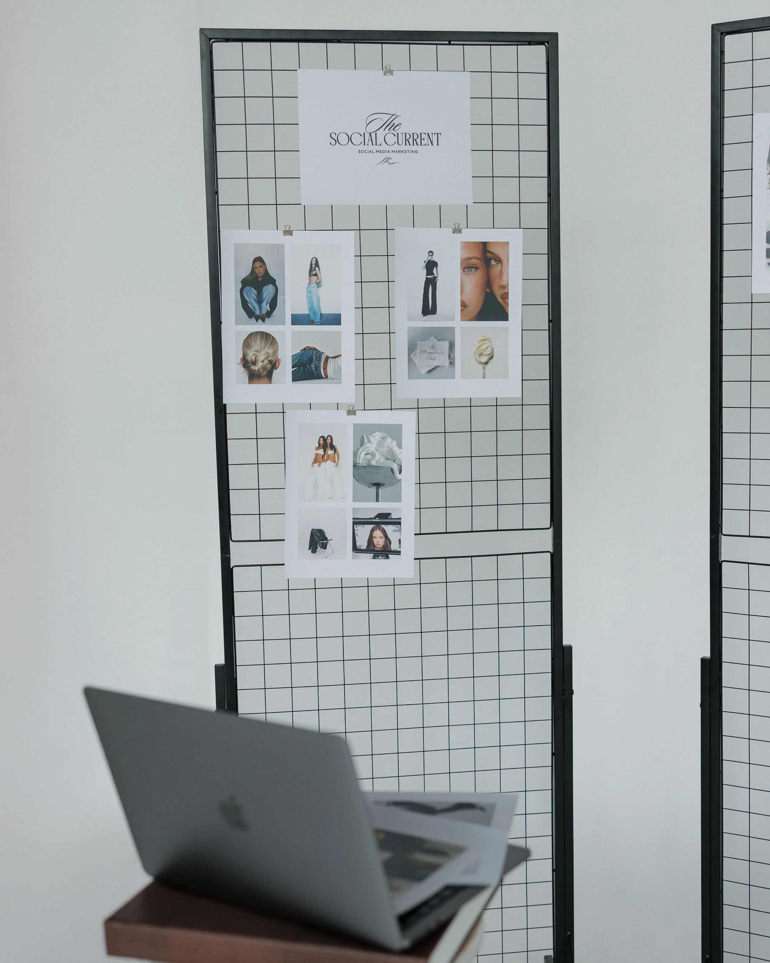 A black wire grid display with photos and a sign titled 'The Social Current' atop, set against a plain white wall, with a silver Apple MacBook on a wooden desk in the foreground.