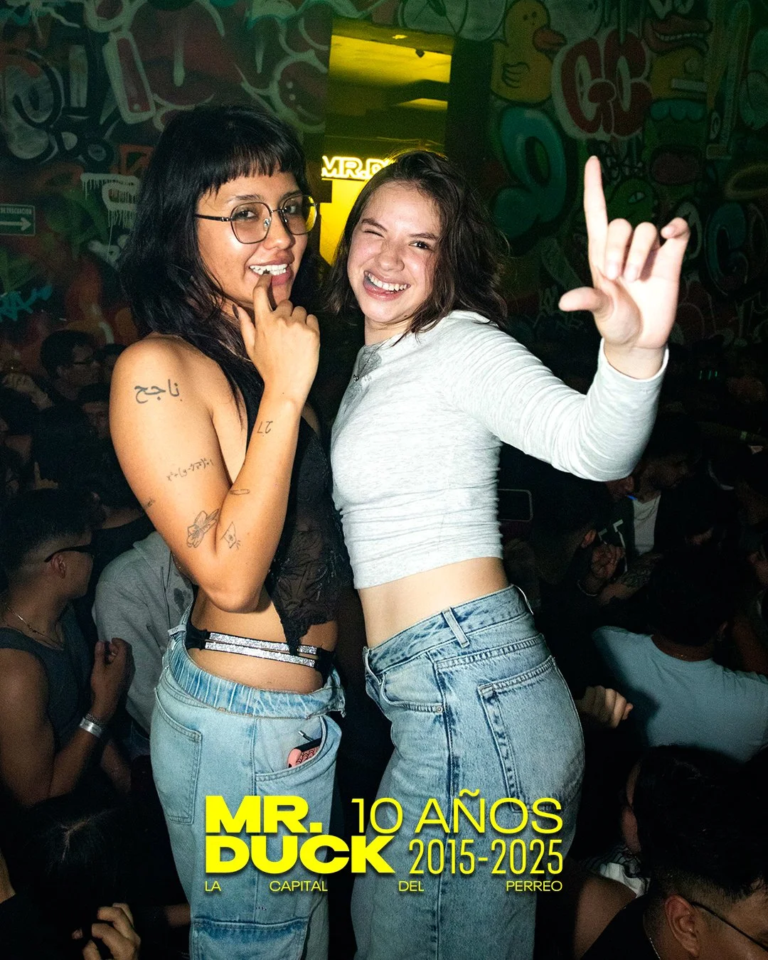 Two women at a crowded party, one with black hair and glasses, the other with brown hair, both smiling and posing for the photo. The background features colorful graffiti-style wall art and dim lighting.