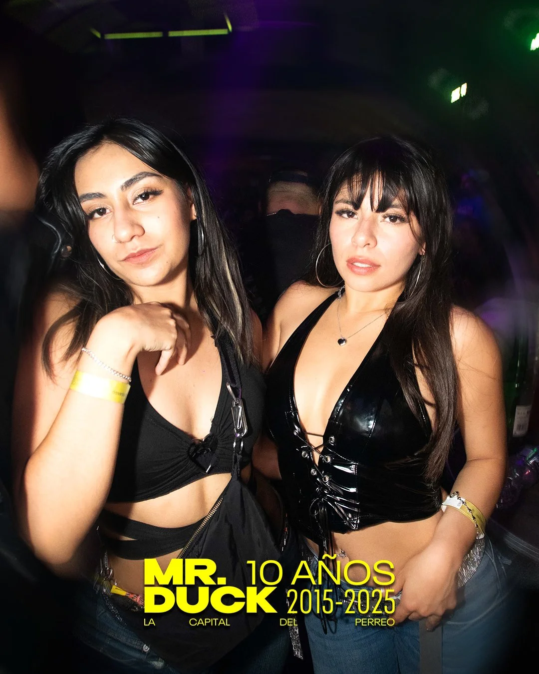 Two women with dark hair and earrings in a nightclub, wearing black tops, posing for a photo with multicolored lights in the background. There is a yellow text overlay that reads 'MR. DUCK 10 AÑOS 2015-2025 LA CAPITAL DEL PERREO.'