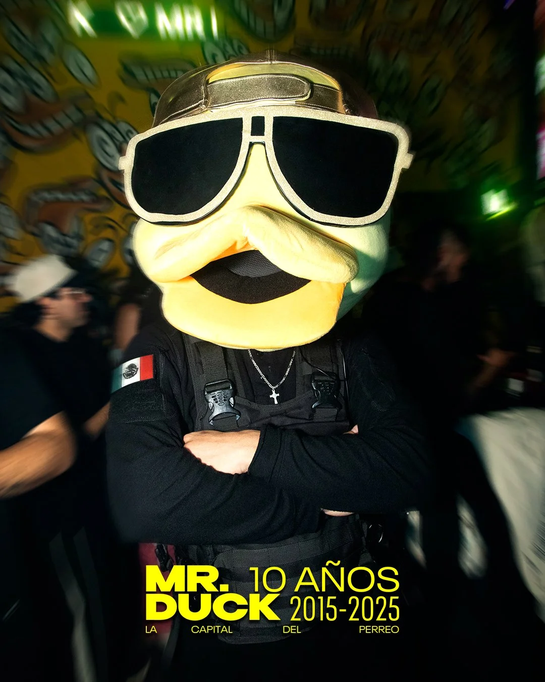 Person wearing a large duck mascot head with sunglasses, crossing arms, in a busy indoor setting, with yellow text at the bottom reading 'MR. DUCK 10 AÑOS 2015-2025, La capital del Perreo'.
