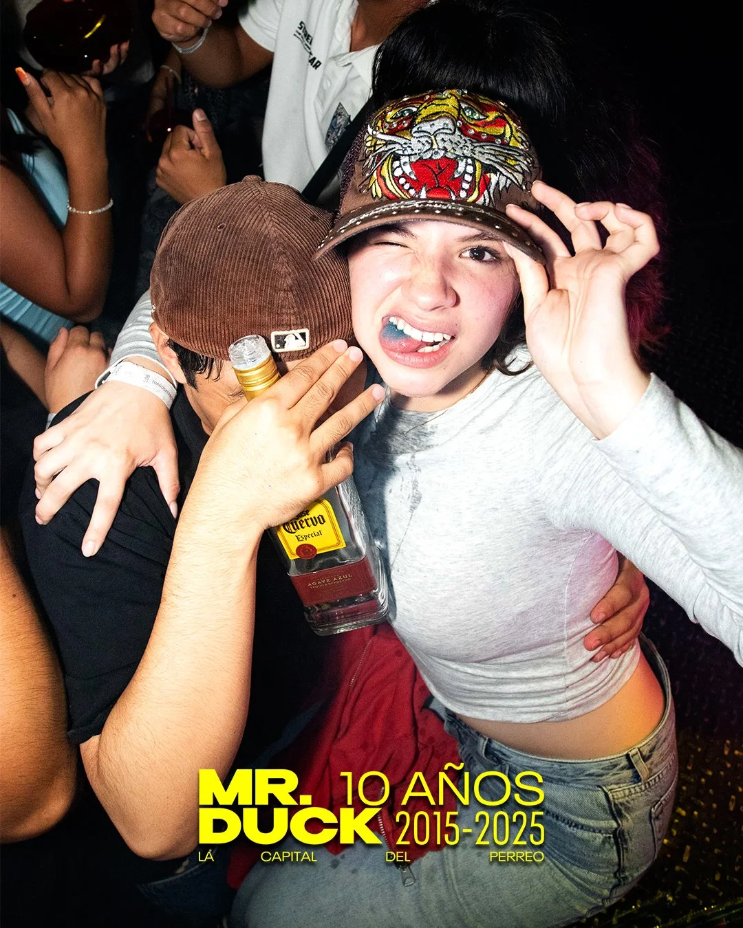 Group of people celebrating at a party, with one woman in a gray shirt and a tiger-themed visor cap making a playful face, holding a bottle of tequila, and surrounded by friends in a lively environment.