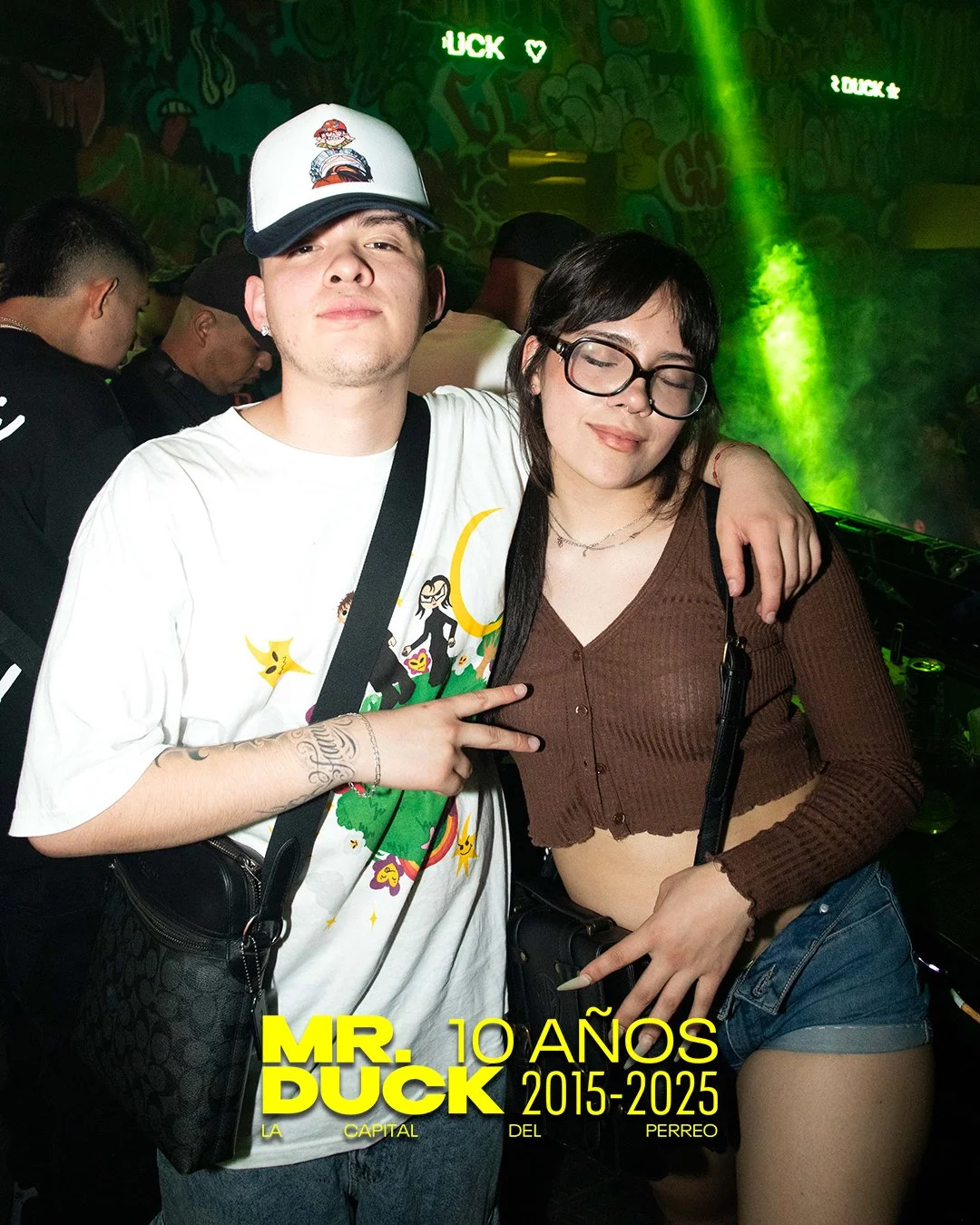 Two young adults, a man and a woman, posing together with a nightclub background and green lighting. The man is wearing a white graphic t-shirt, a white and black cap, and has tattoos on his arm. The woman is wearing glasses, a brown crop top, and de