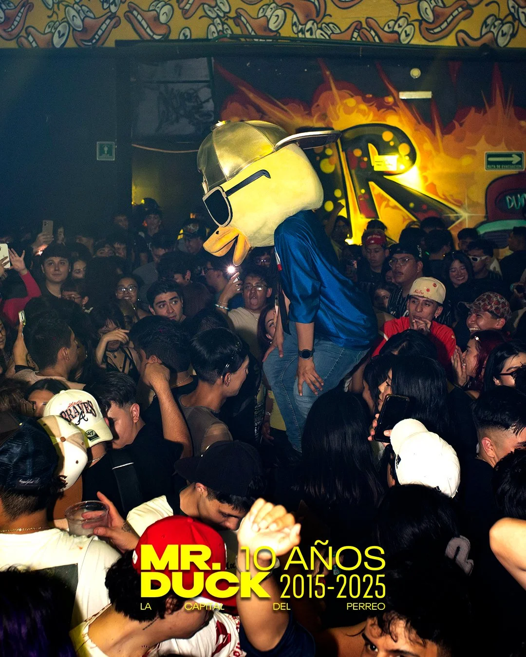 A crowded indoor party with a person wearing a large duck mascot head, standing on others' shoulders, wearing a blue sports jacket. The background features graffiti-style wall art with vibrant colors and a crowd of young people, some taking photos, a