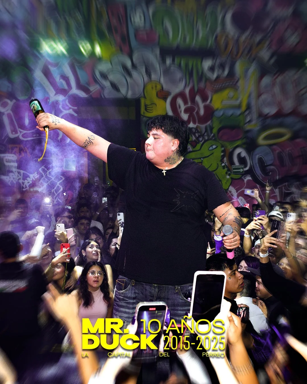 A performer on stage holding a drink and microphone in a crowded nightclub with graffiti art on the wall in the background. Audience members are taking photos and videos.