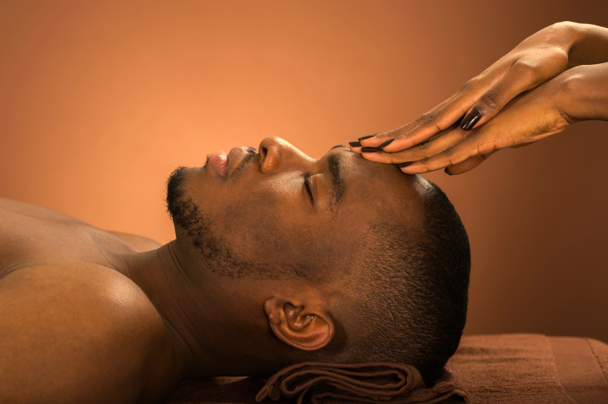 A man lying on a massage table with eyes closed, receiving a facial massage from a therapist's hands.