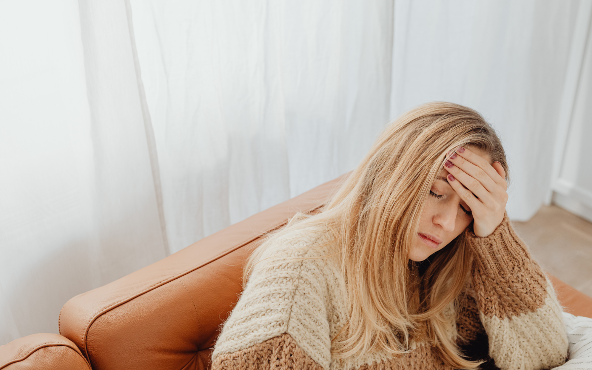 A woman sitting on a tan leather couch, holding her forehead with her hand, appearing to have a headache.