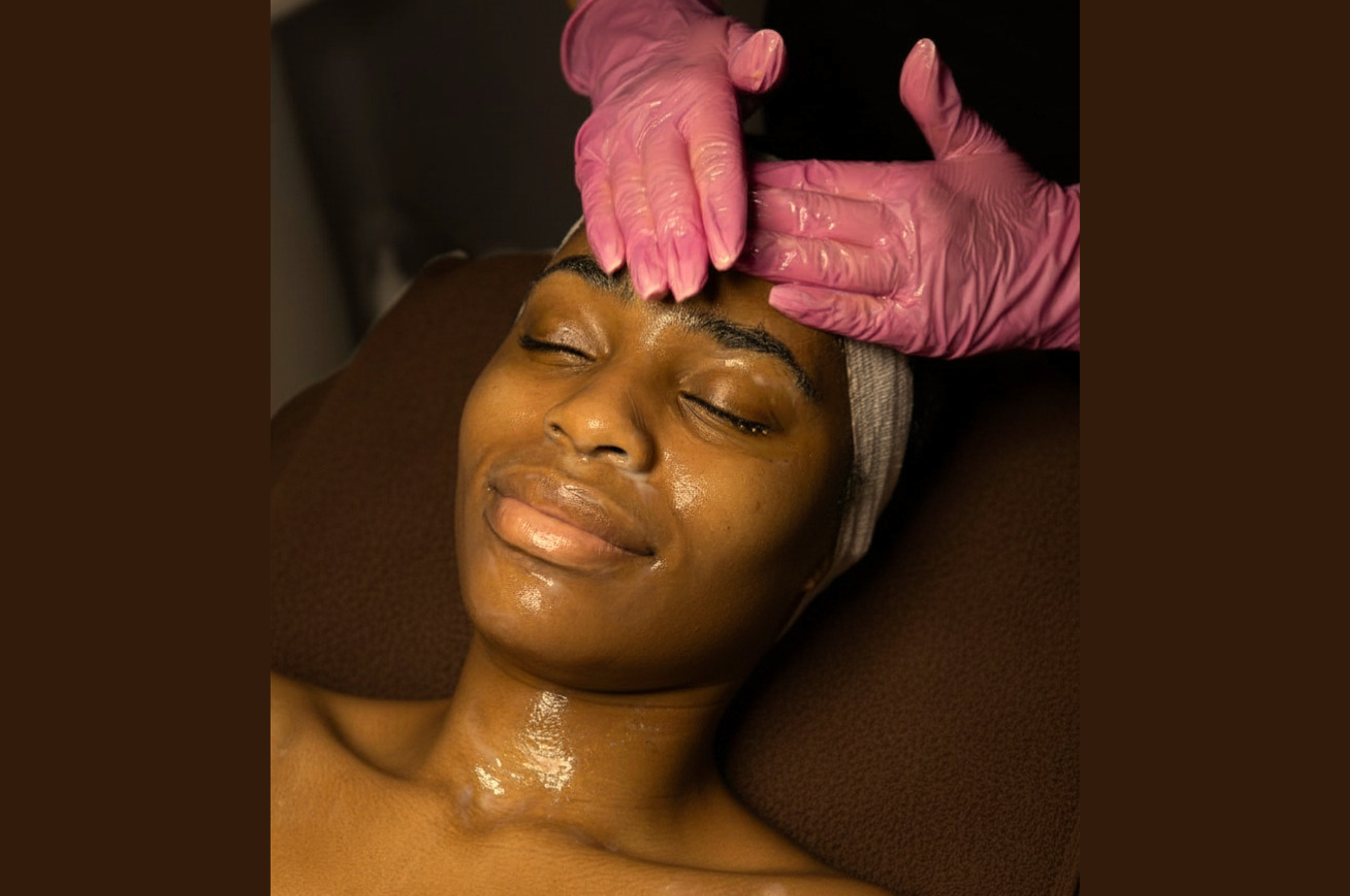 A woman receiving a facial treatment, lying on a brown spa bed, with her eyes closed, a relaxed smile, and a towel wrapped around her head. A practitioner wearing pink gloves is gently applying a product to her forehead.