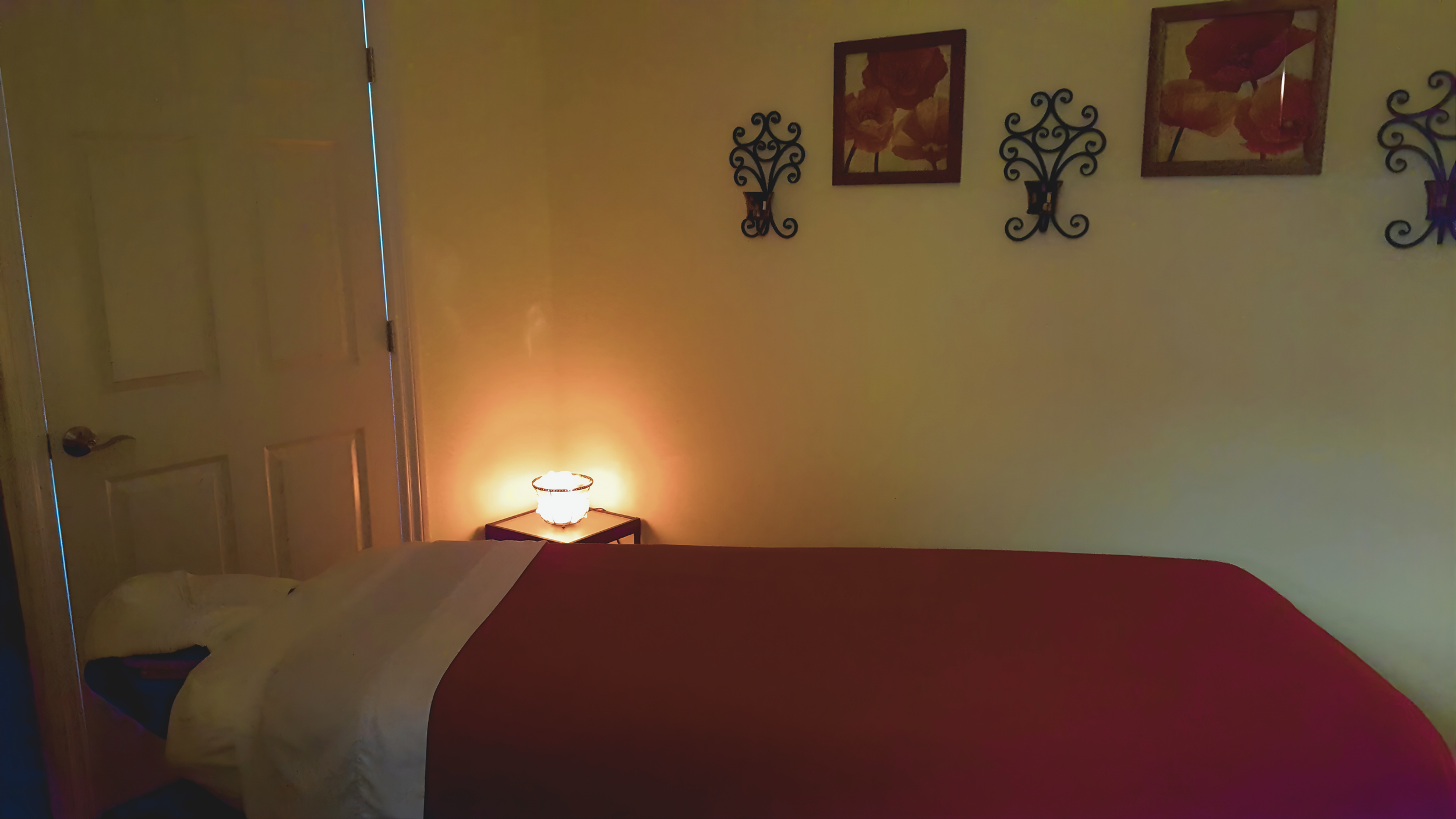 A bedroom with a bed covered in a red blanket, pillow, and a yellow blanket, a nightstand with a lit lamp, and three framed floral pictures with black decorative wall sconces on a yellow wall.