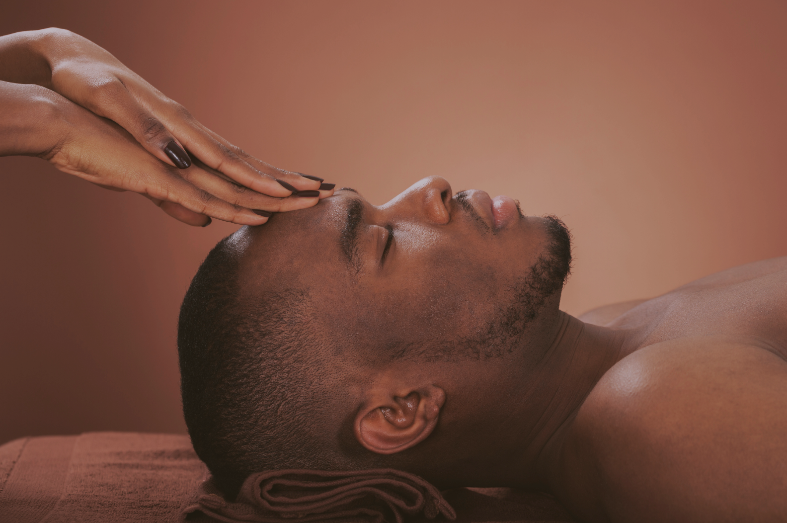 A man is lying down with his eyes closed, receiving a forehead massage from a person with dark nail polish in a spa setting.