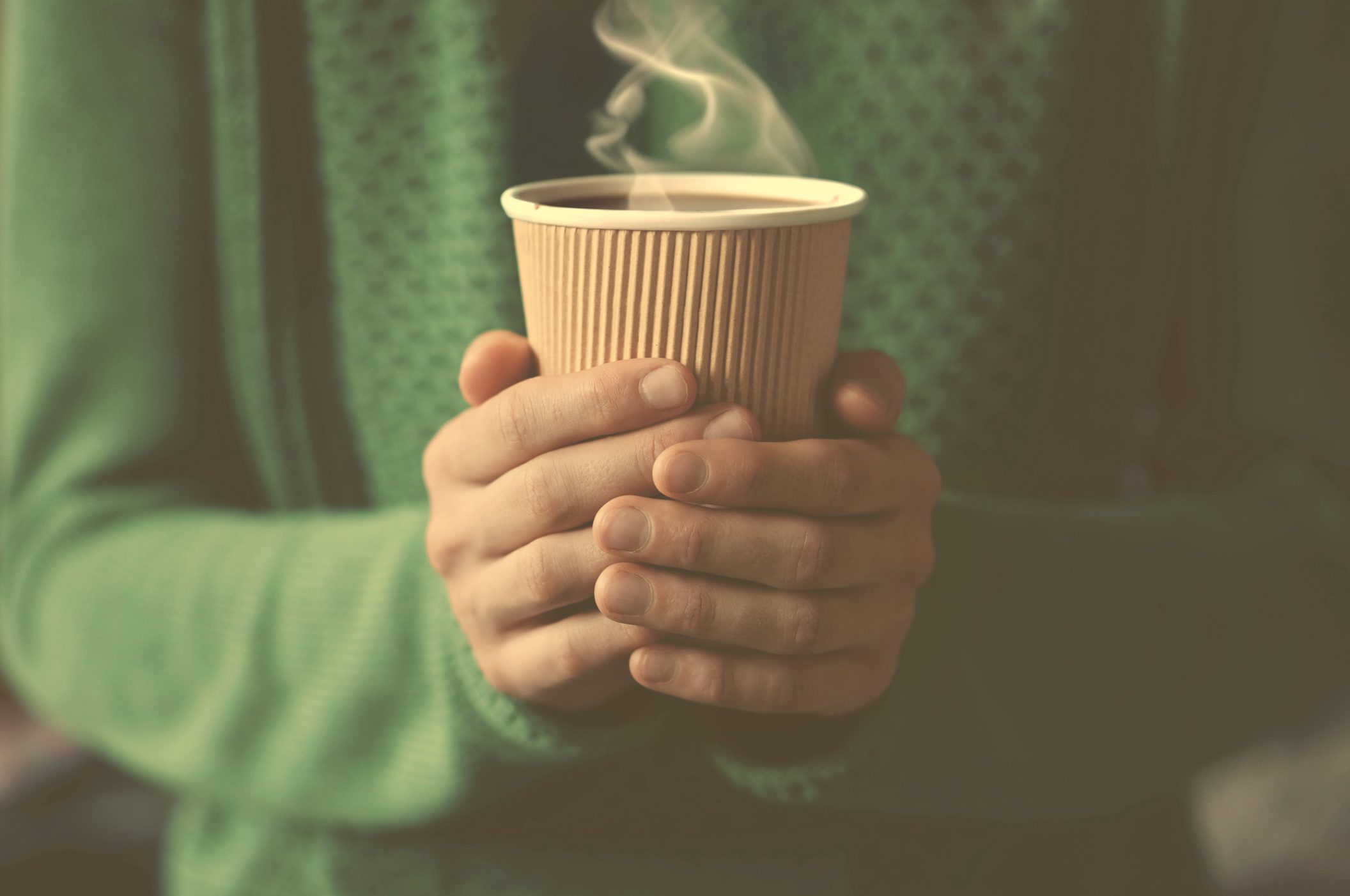 Person wearing green sweater holding a paper coffee cup with steam rising from it.