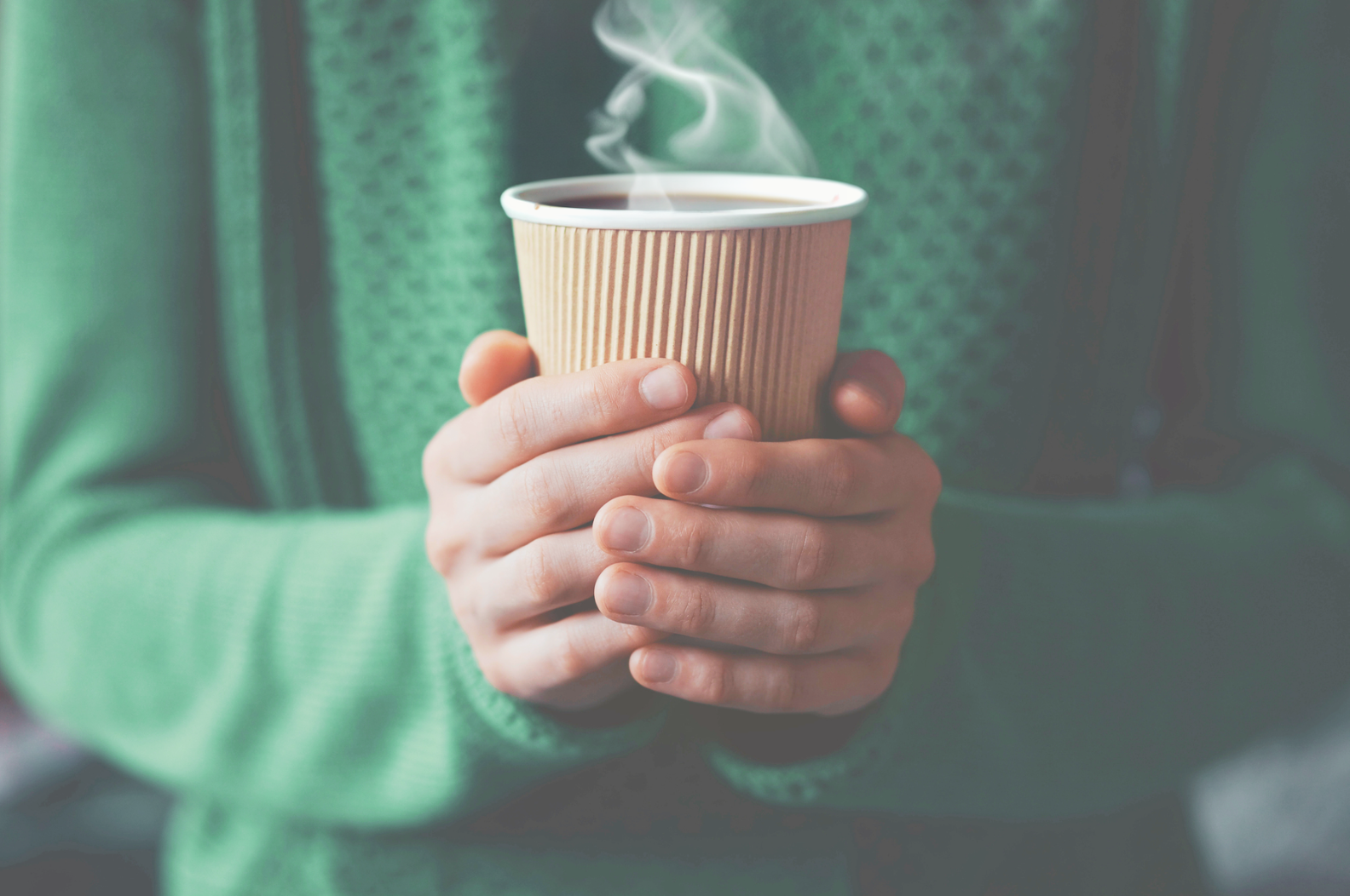 Person holding a steaming cup of hot coffee or tea in a brown corrugated paper cup with a green sweater.