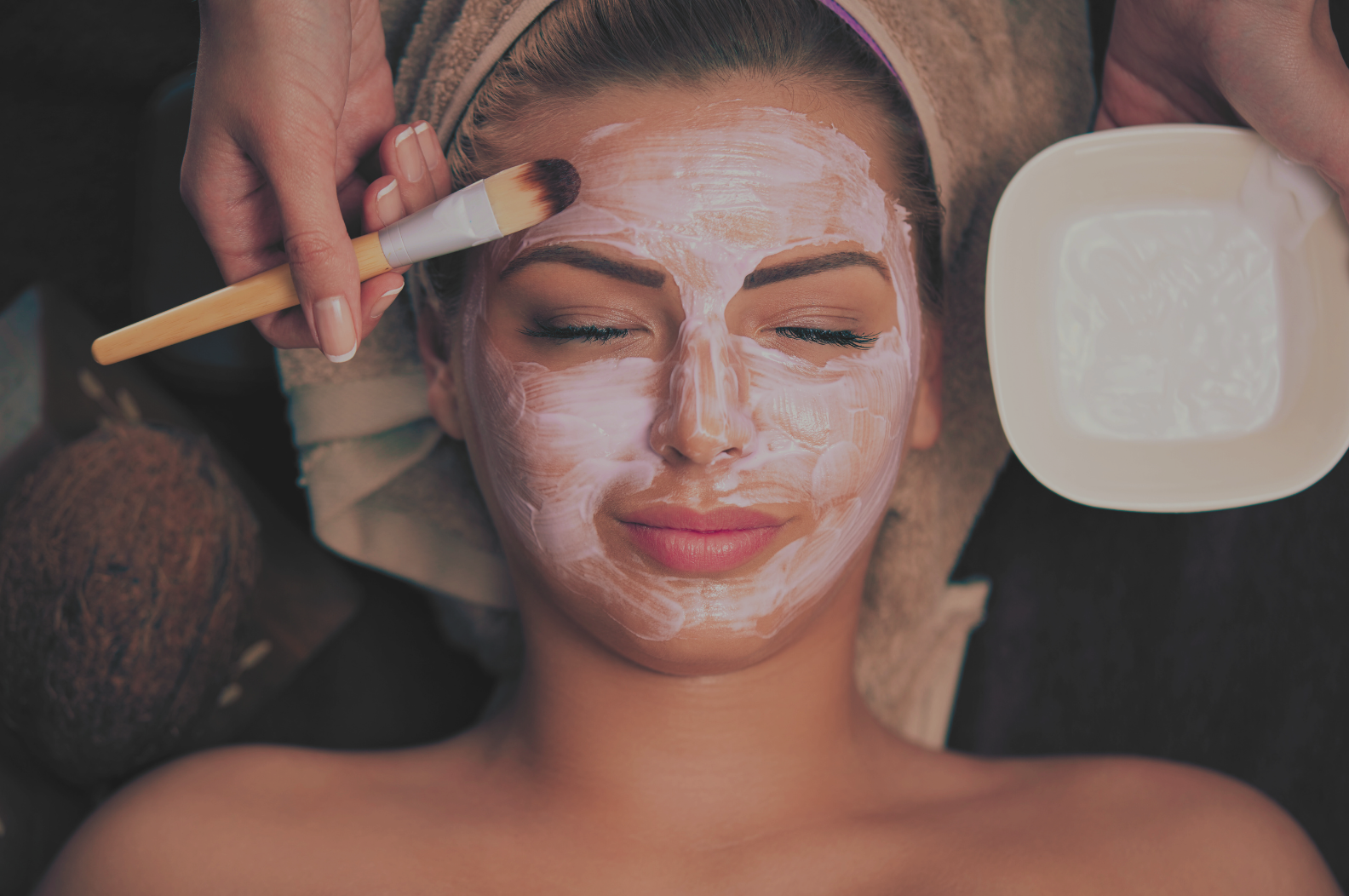 A woman receiving a facial treatment, with her eyes closed, as a skincare professional applies a facial mask with a brush.