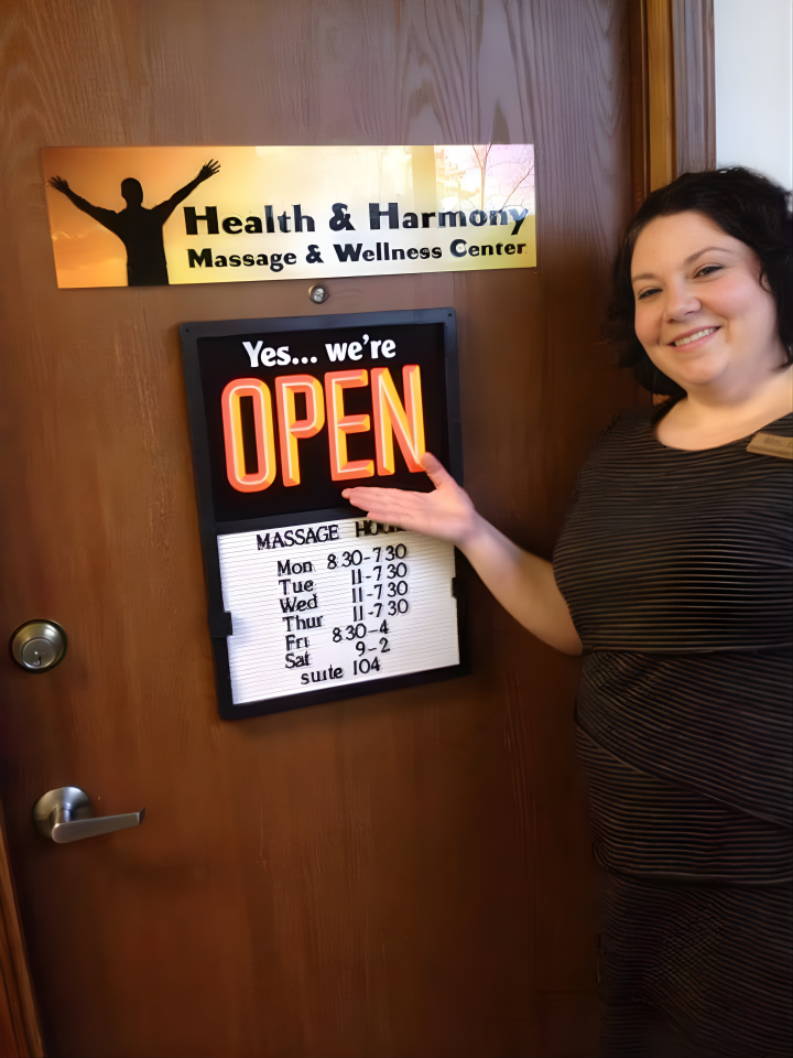 A woman standing next to a sign on a wooden door for a health and wellness center, smiling and gesturing towards the open sign. The sign indicates the center is open, with hours listed for Monday through Saturday.
