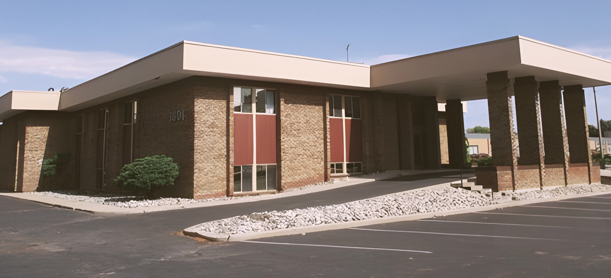 A mid-century modern commercial building with brick walls, large rectangular windows with maroon accents, a flat white roof extending over the entrance supported by brick columns, and a parking lot with marked spaces and landscaped areas with rocks and bushes.