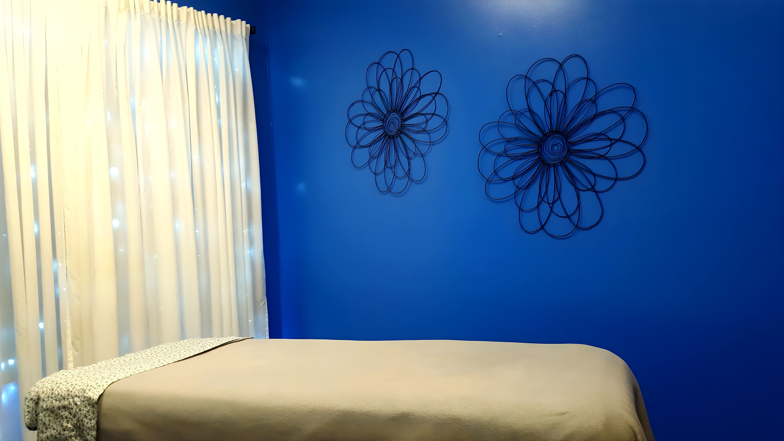 A bedroom with a blue wall decorated with two black wire flower-shaped wall art pieces, white curtains with small lights, and a bed with beige bedding and a patterned blanket at the foot.
