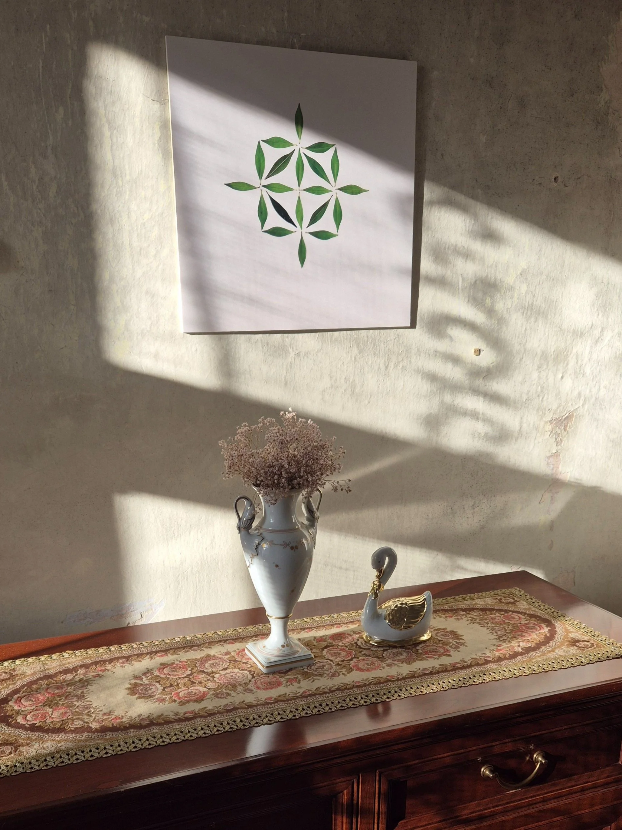 A wooden sideboard with a floral-patterned runner, a white ceramic vase with pink flowers, and a ceramic swan figurine with gold accents, against a textured wall with a geometric artwork shadow.