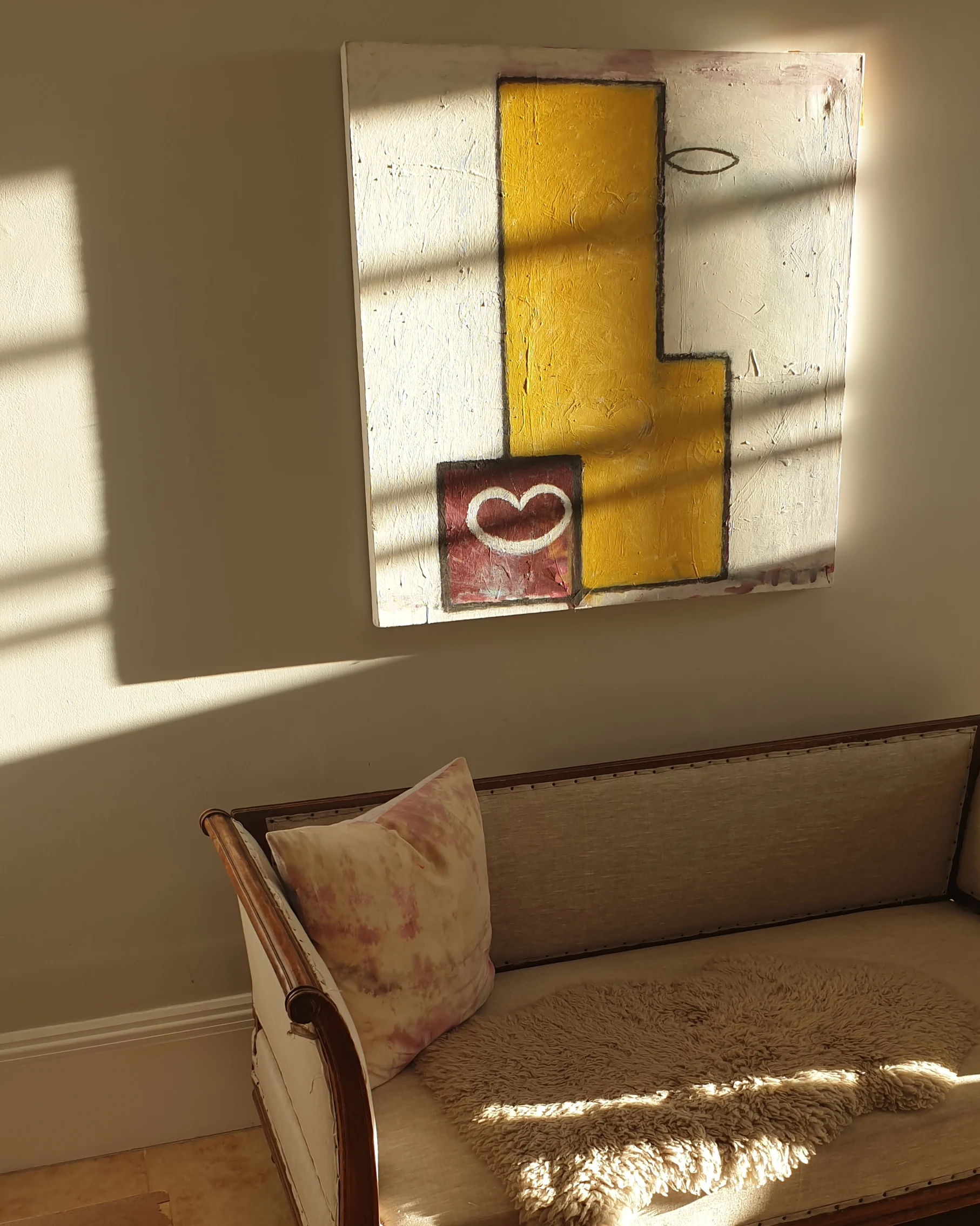 A colorful abstract painting with yellow, white, and red shapes hangs on a beige wall. A shadow from window blinds falls across the painting. Below, a vintage cream-colored sofa with a pink tie-dye pillow and a beige fur throw is seen.