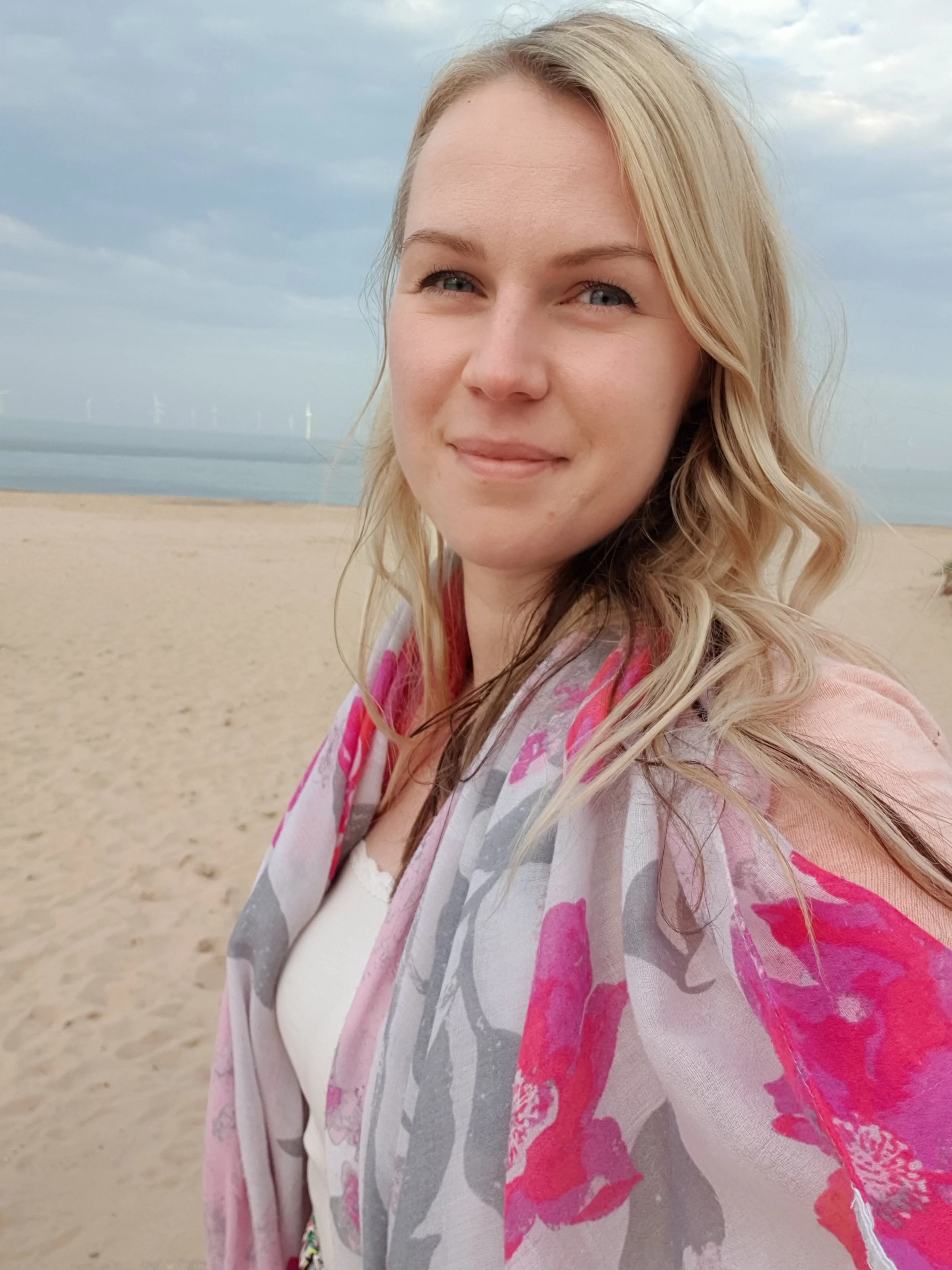 A woman with blonde hair standing on a sandy beach with wind turbines in the ocean in the background, under a partly cloudy sky.