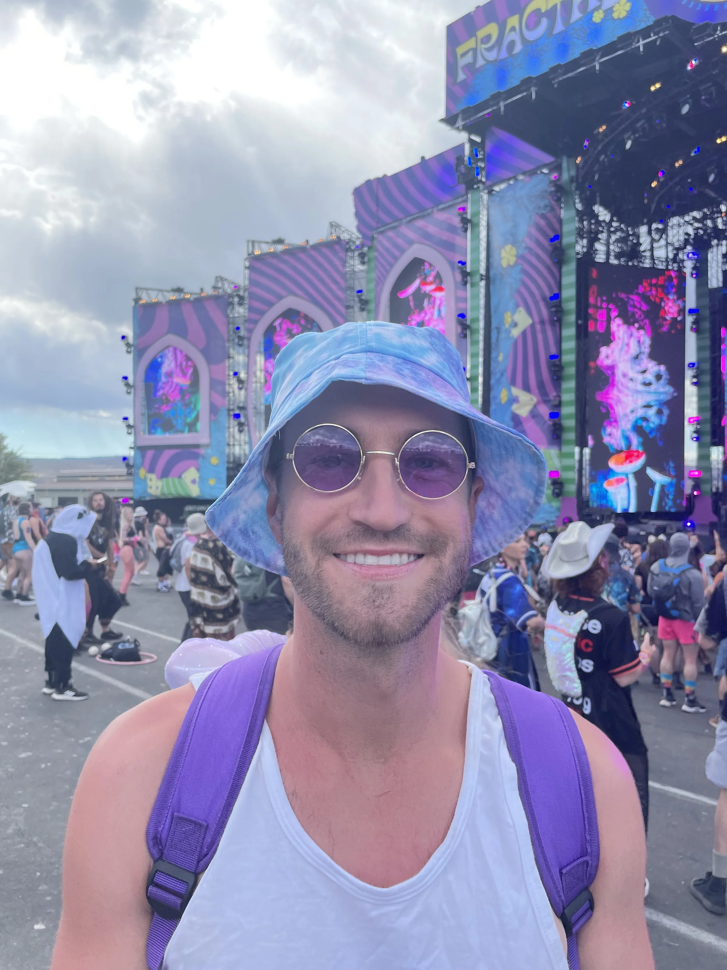 Person in a festival crowd wearing a tie-dye bucket hat, round sunglasses, white tank top, and purple backpack, in front of a colorful stage with large screens and decorative panels.