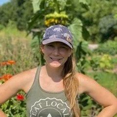 Young woman, Briana Cushman, instructor at Bluff Strokes Art Center, wearing a patterned baseball cap and olive green tank top, standing outdoors in a garden with flowers and greenery.