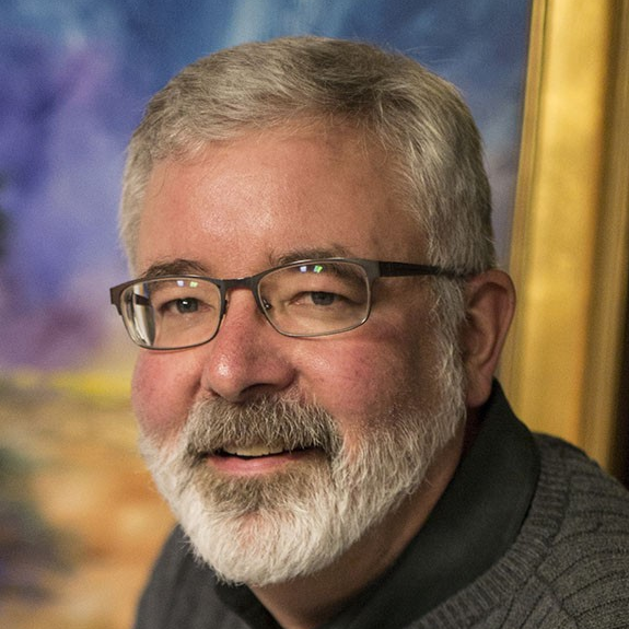 Close-up of a middle-aged man with glasses, gray hair, and a beard, smiling indoors with a colorful background.