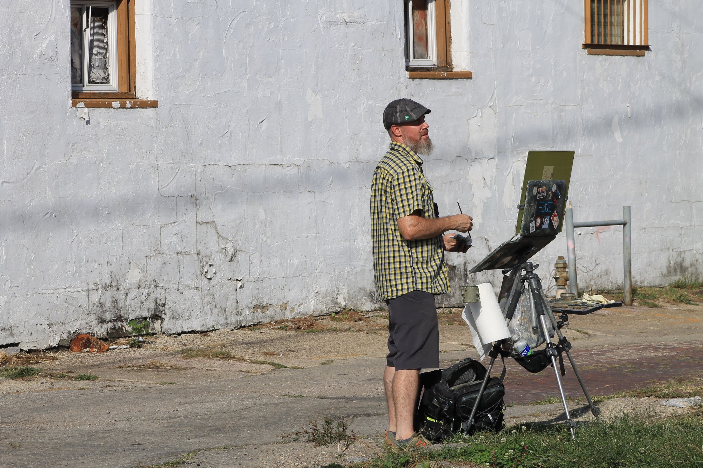 A person sitting on sidewalk, painting on a small wooden box, wearing a straw hat, blue striped shirt, and jeans, with a wheelchair nearby.