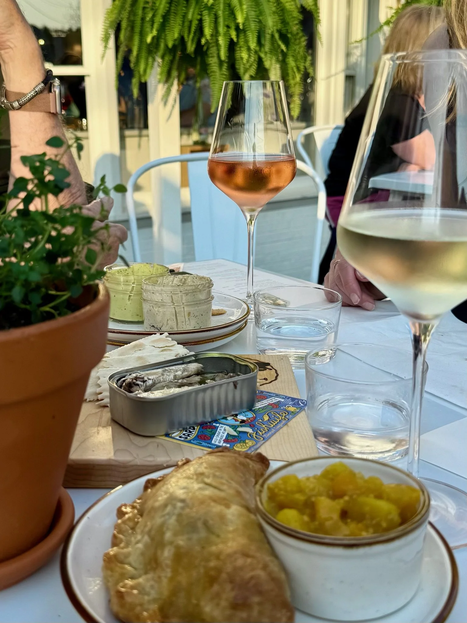 Table setting with two glasses of white wine and a rosé wine, a plate with a pastry and a small bowl of yellow fruit, a can of sardines, and some bread or spread, outdoors with greenery in the background.