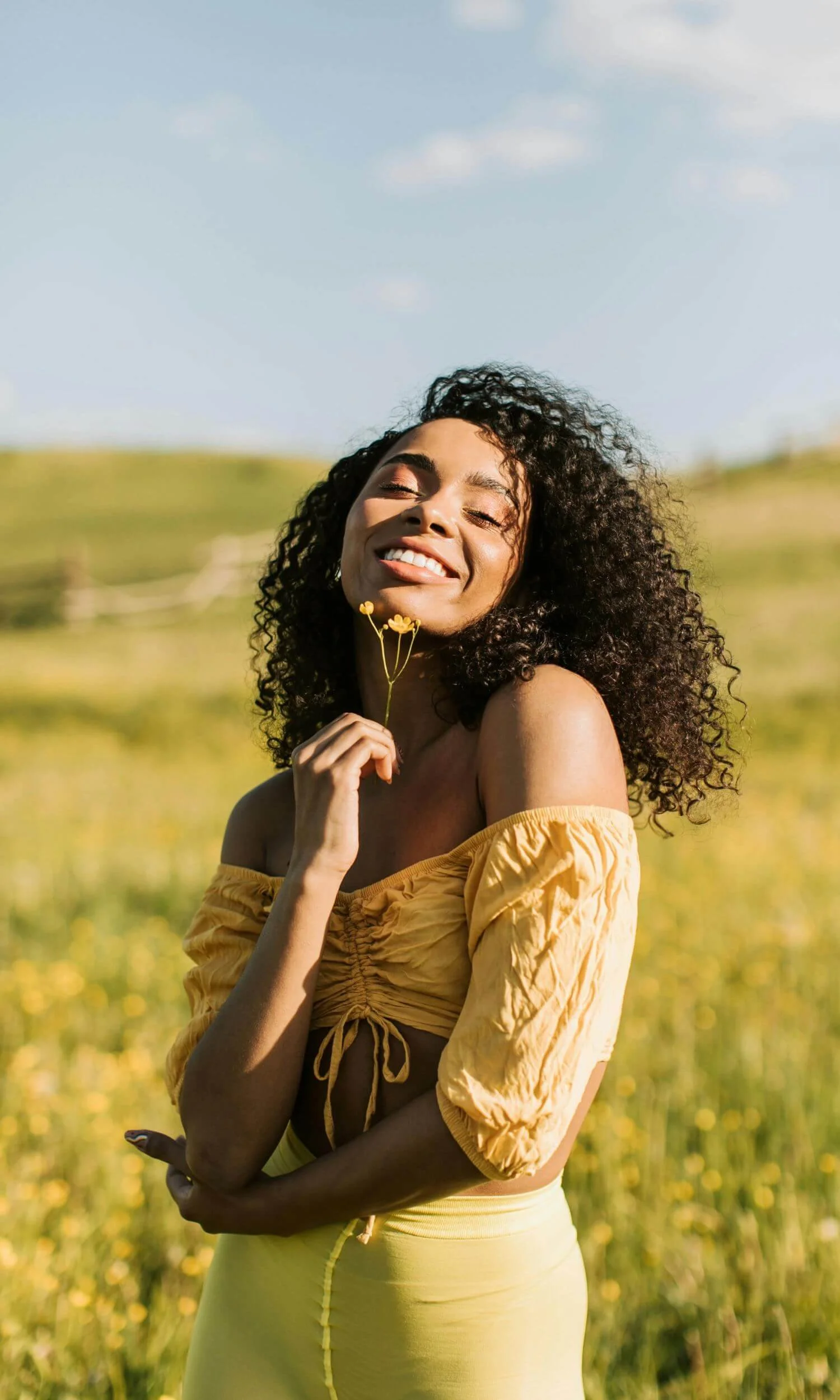 Black woman with curly hair holding a flower and smiling in a yellow field