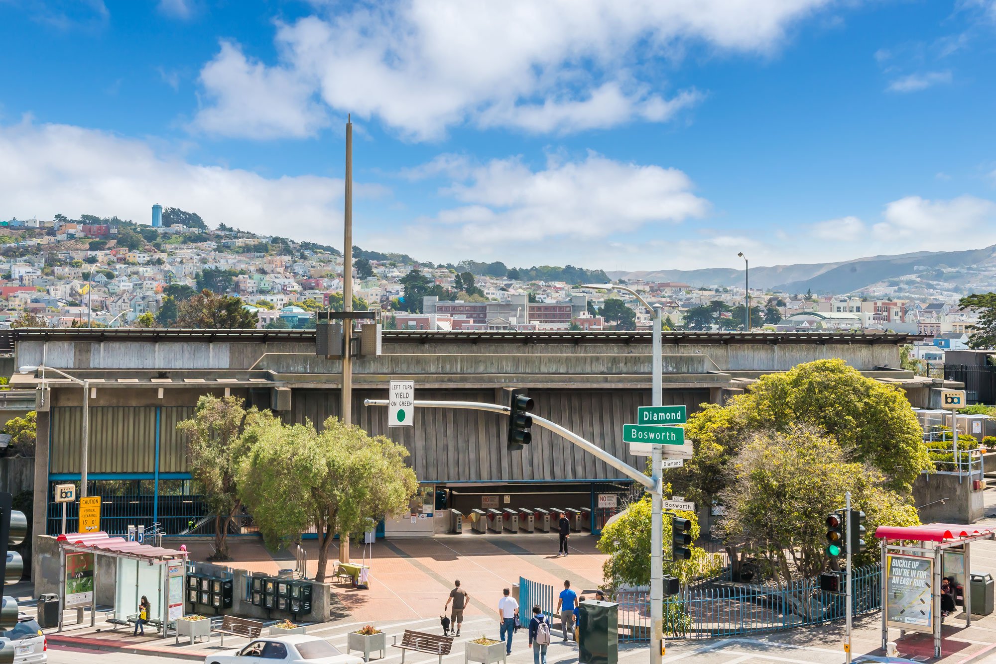 View of an urban train station with trees, directional signs for Diamond and Bosworth, and a cityscape with colorful houses on a hillside in the background under a partly cloudy sky.