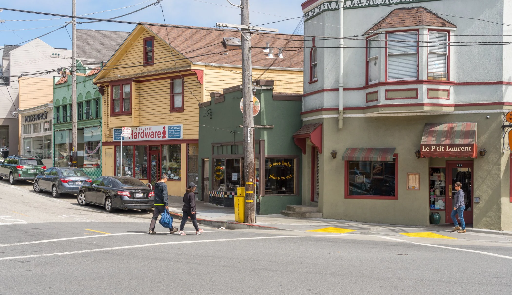 A street view with colorful buildings, parked cars, and pedestrians crossing the road. Shops include a hardware store and a restaurant named Le P'tit Laurent.