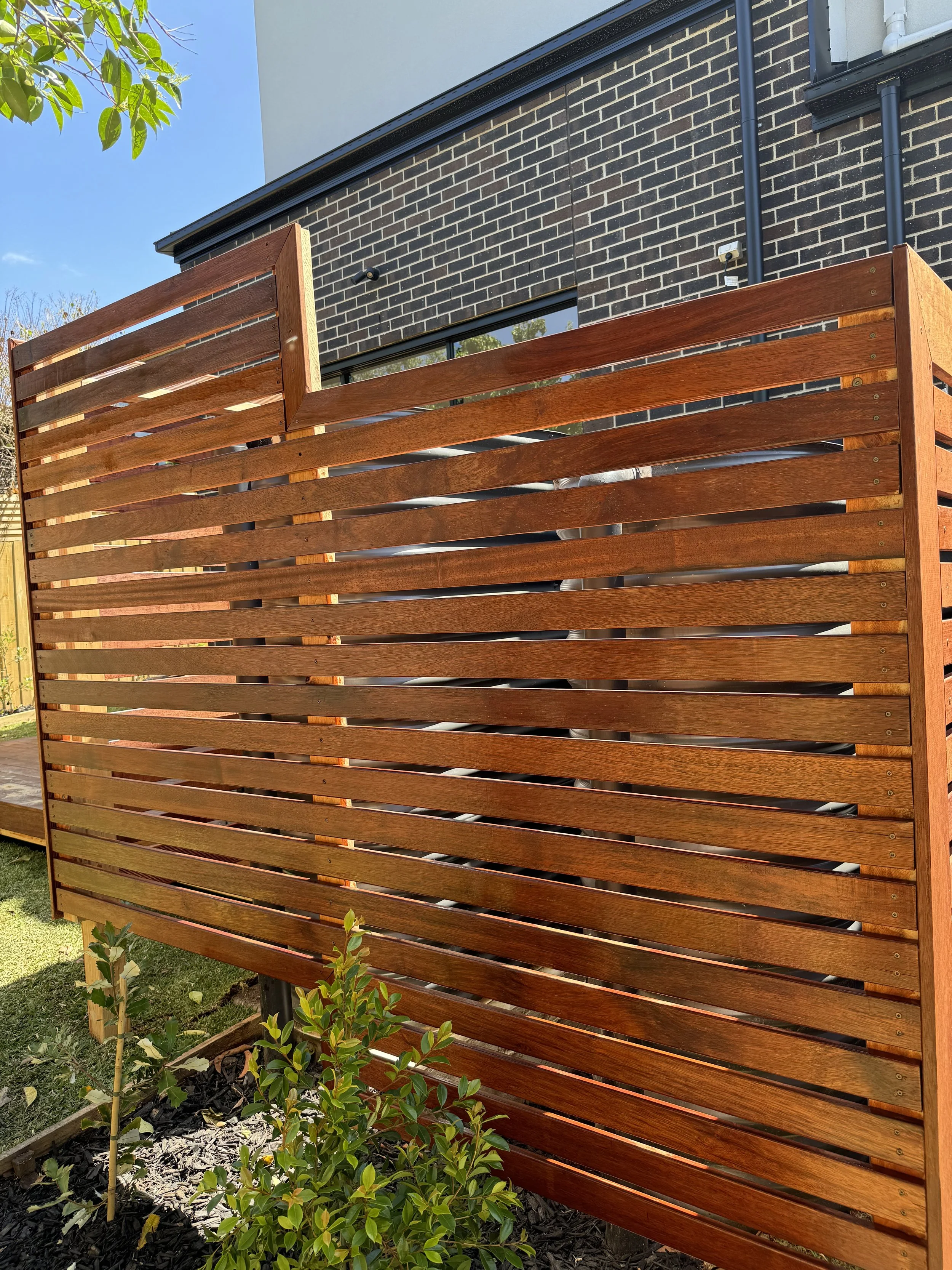 A wooden privacy screen with horizontal slats installed in a backyard garden, with a brick house and some green plants nearby.