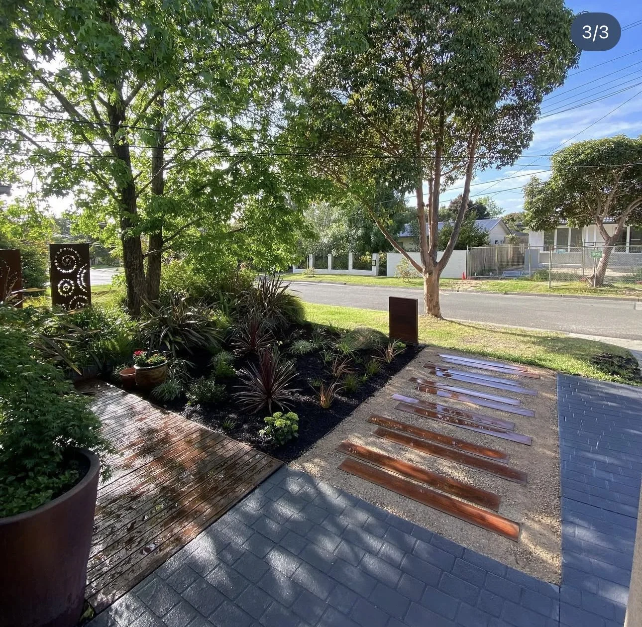 A front yard with a garden bed of dark brown soil and various plants, including ferns and grass plants, bordered by a wet wooden deck and a gray brick pathway. Two metallic decorative panels and a black sign are also present, with a large tree provid