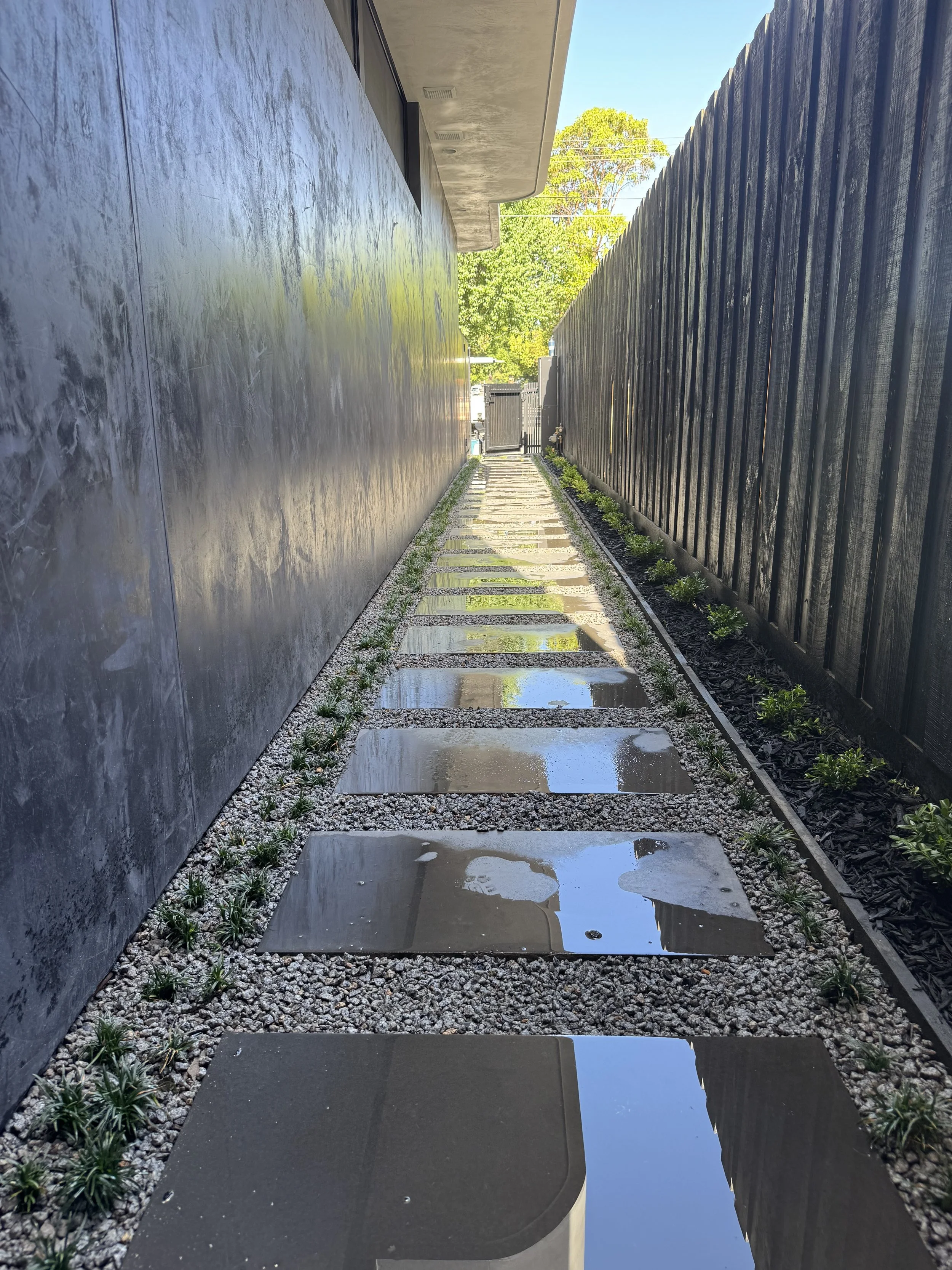 A modern outdoor pathway with large, flat, soaking wet tiles, flanked by a stone gravel border with small plants, leading to a gate on the horizon, with a building on the left and a tall dark fence on the right, under a clear blue sky.