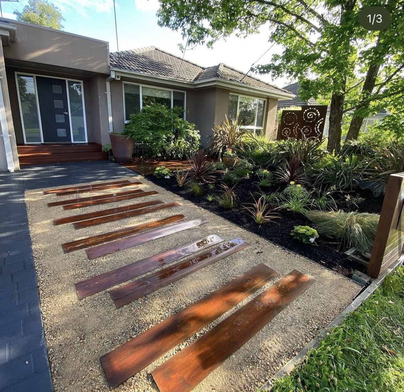 A front yard with a sidewalk made of wooden planks and gravel, leading to the house's entrance. The house has large windows and a door with modern design, surrounded by lush green plants and trees. A decorative metal panel with spiral patterns is vis