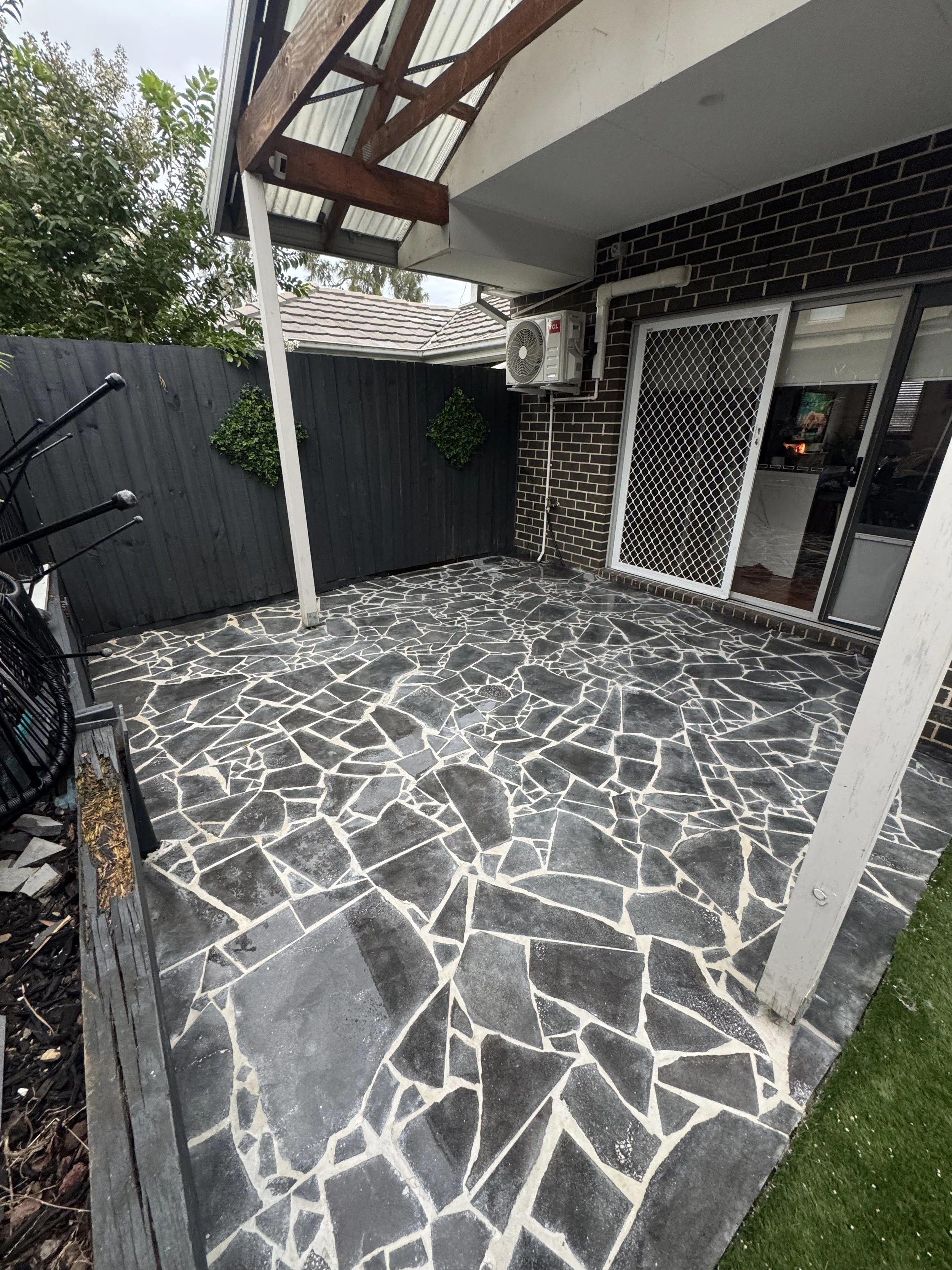 Small outdoor patio with a decorative stone floor, black wooden fence with greenery, sliding glass door with security screen, and an air conditioning unit on the brick wall.
