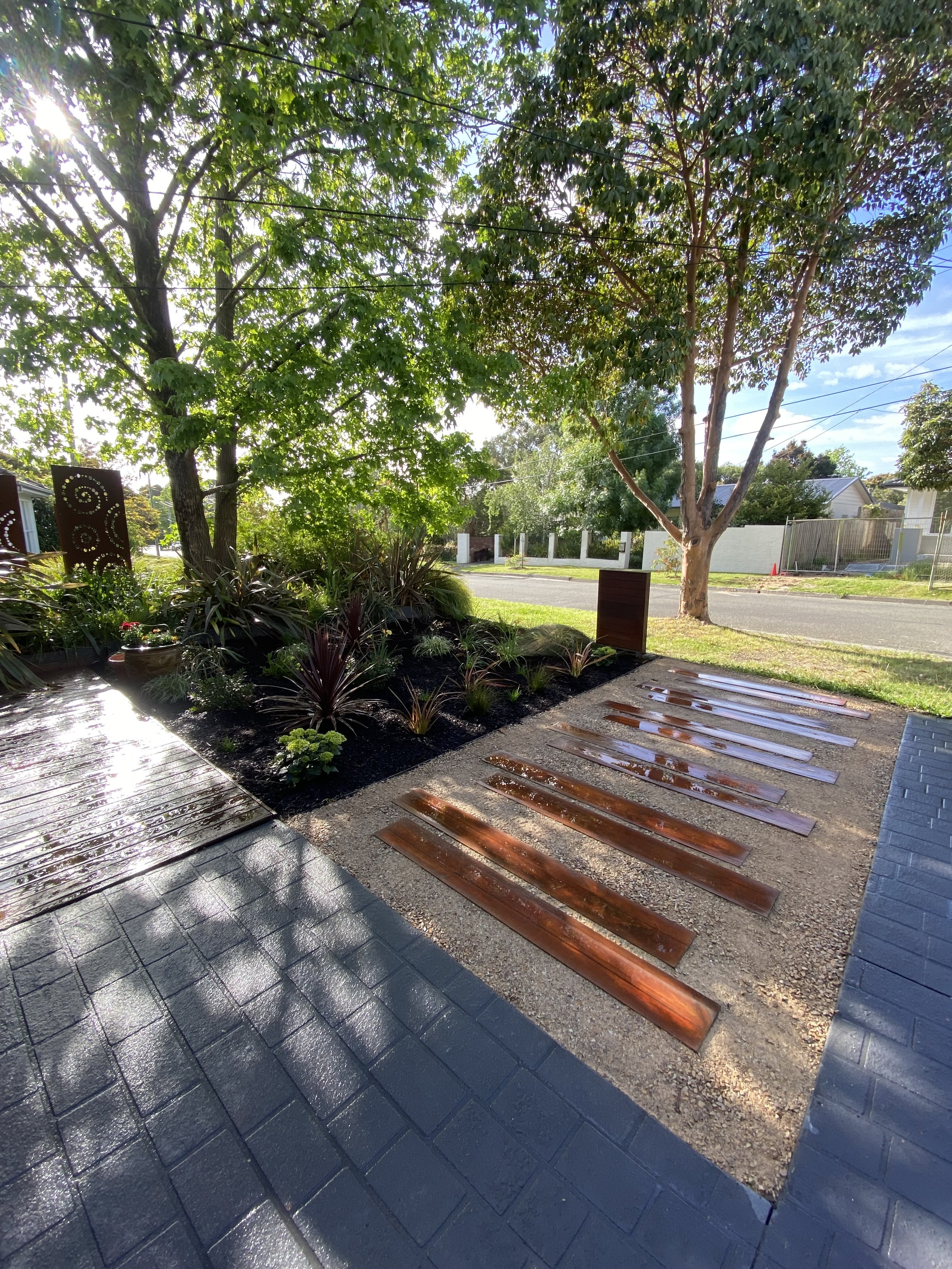 View of a sidewalk with wooden planks laid out on gravel, a landscaped yard with trees and plants, and residential houses across the street on a sunny day.