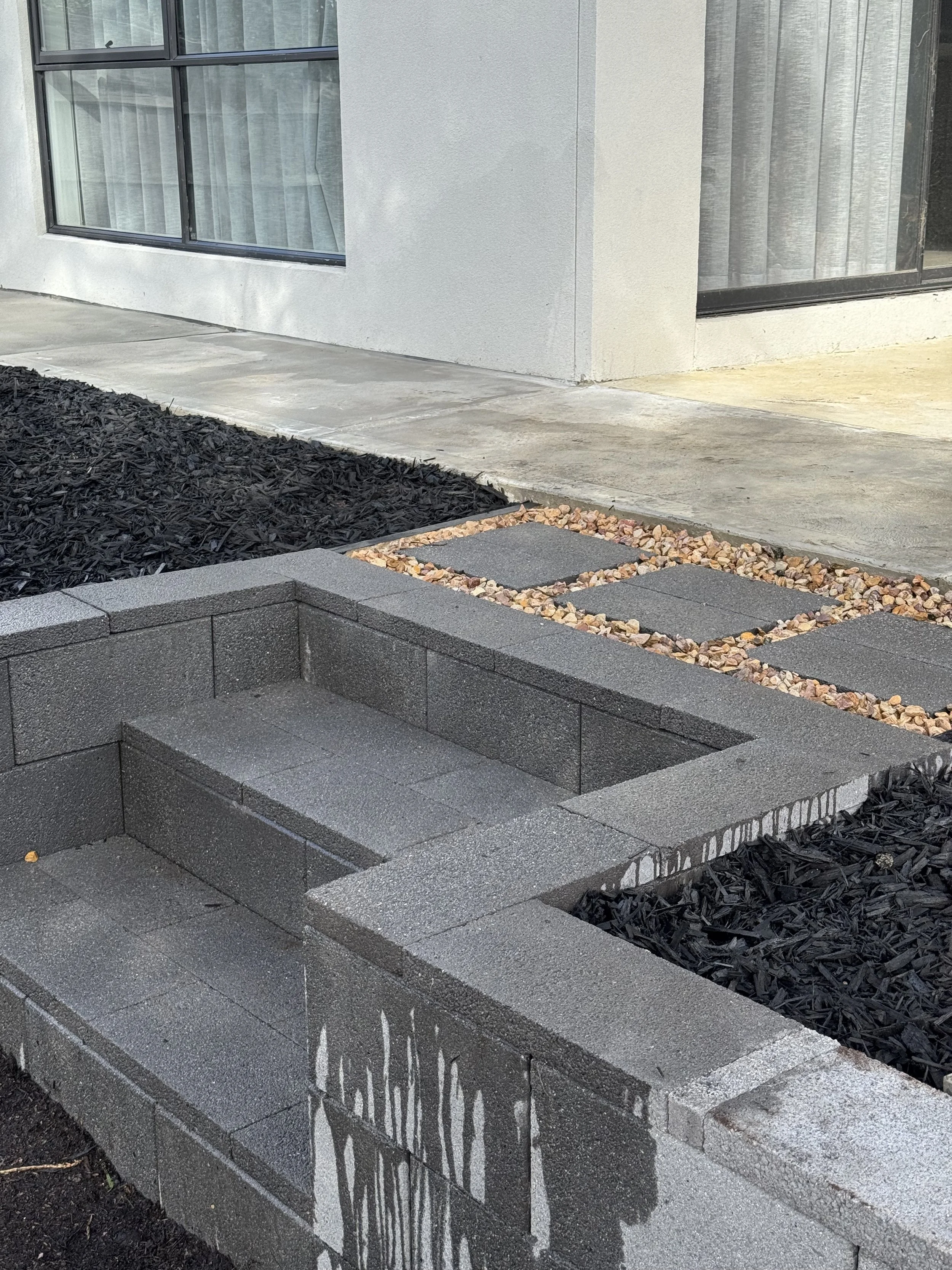 A newly constructed outdoor space with gray concrete pavers, a small raised garden bed made of gray cinder blocks, and a pathway with square stepping stones surrounded by small multicolored rocks, in front of a modern house with large windows and tan