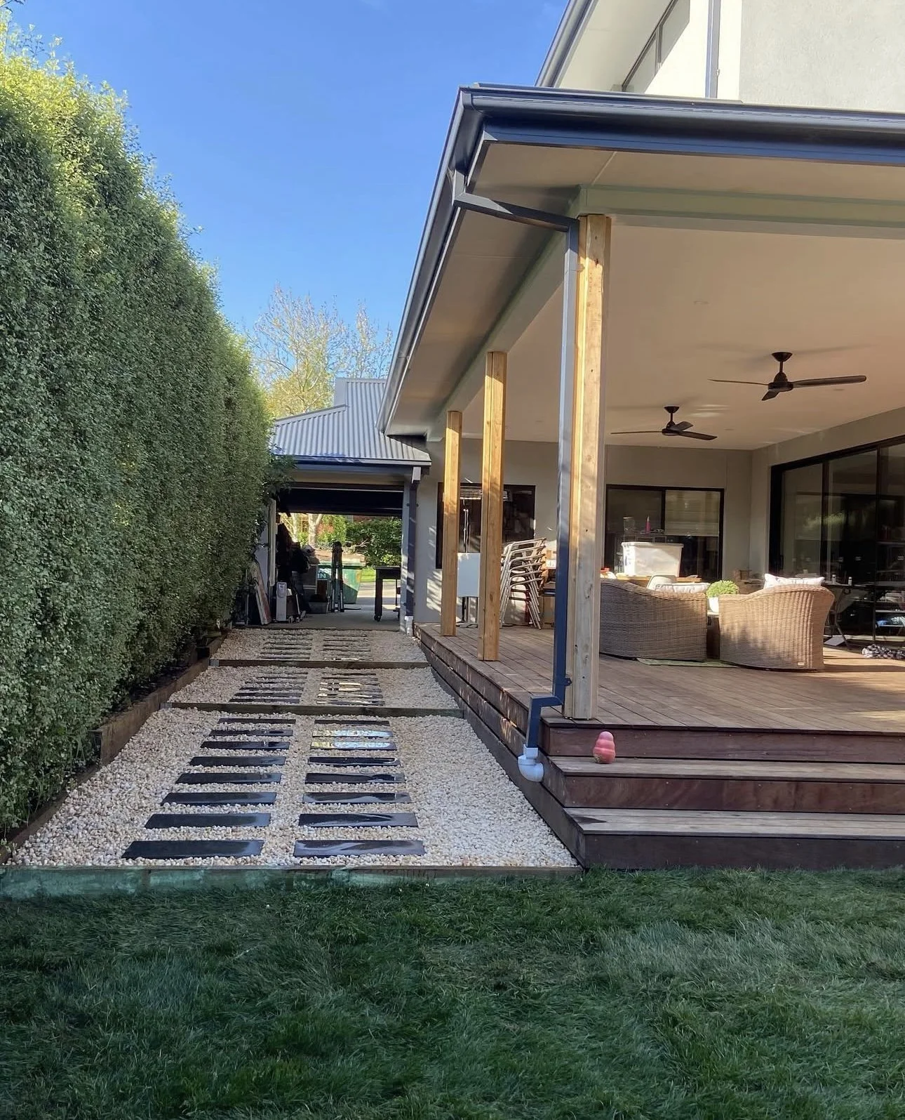 Backyard patio with wooden deck and outdoor furniture, adjacent to a gravel pathway with stepping stones and a tall hedge on the left side.