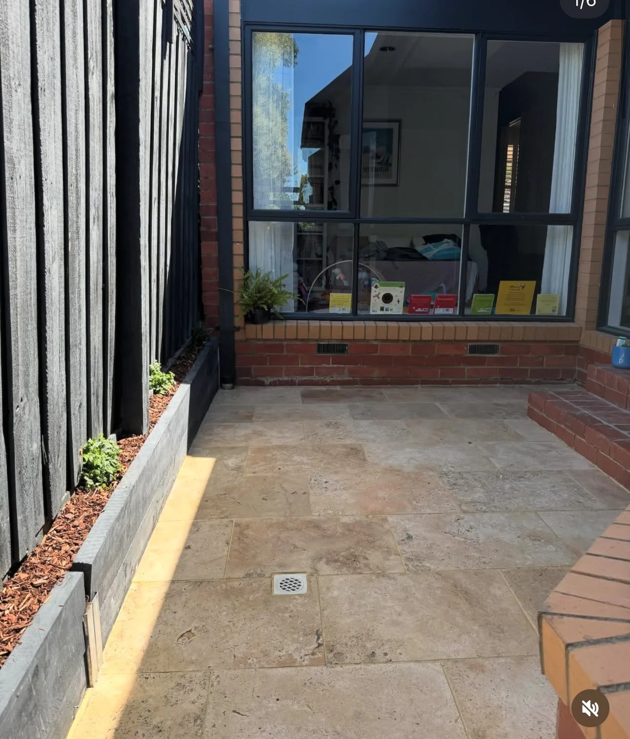 Small patio area with stone tile flooring, a black wooden fence with plants, a large window showing an indoor room with various items, brick walls, and steps leading to another part of the patio.