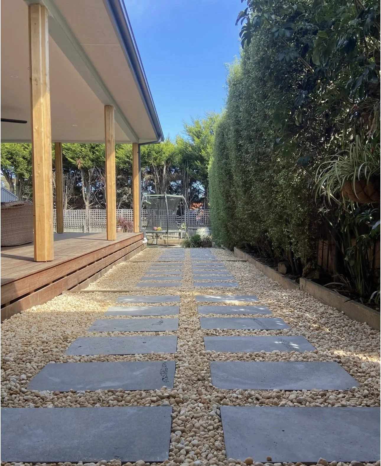 A backyard path with rectangular stepping stones set in gravel, leading to a small patio with a trampoline in the background, surrounded by trees and a tall hedge on the right side.