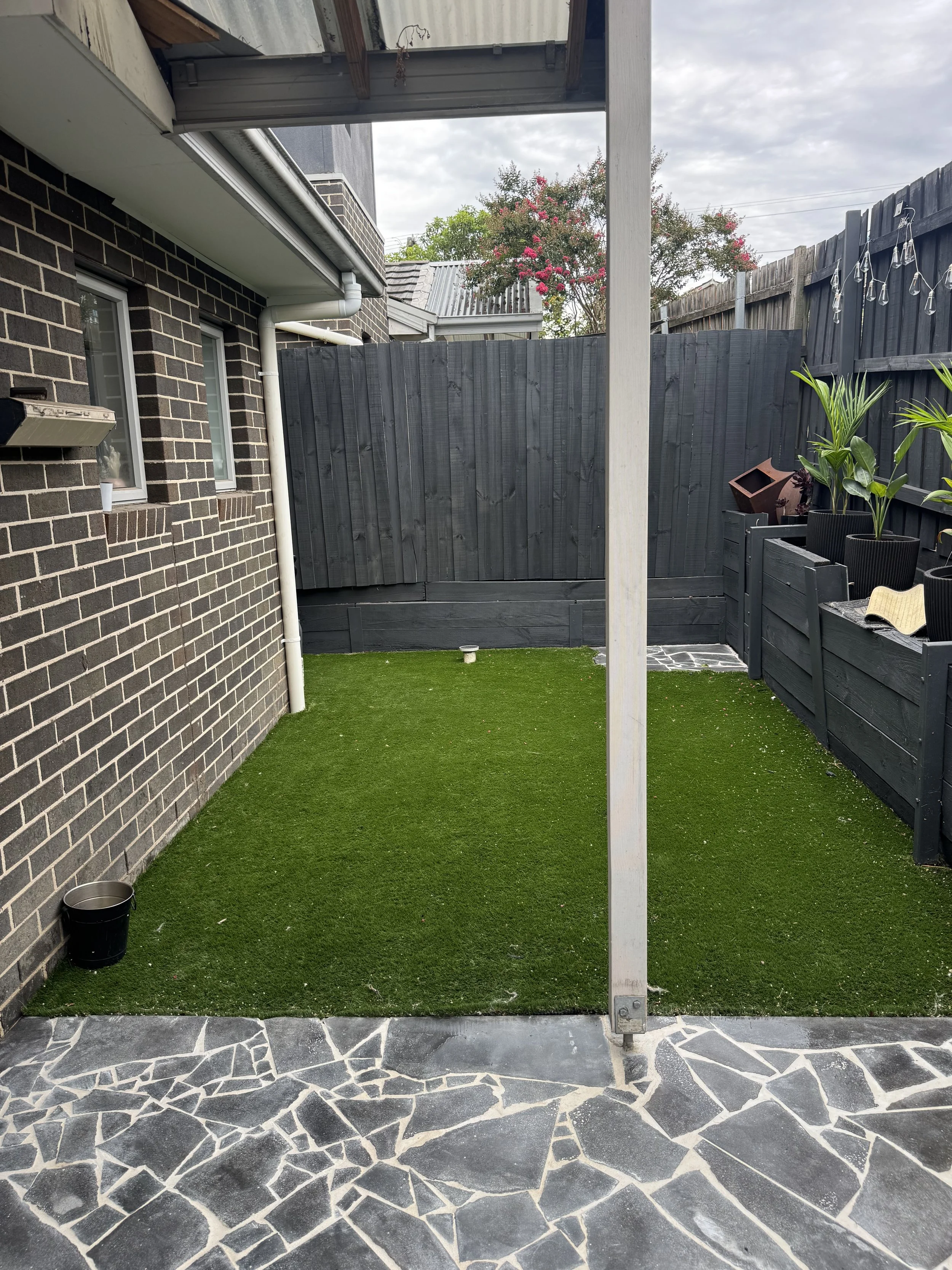 Backyard area with artificial grass, black wooden fencing, potted plants, and stone patio in a residential setting