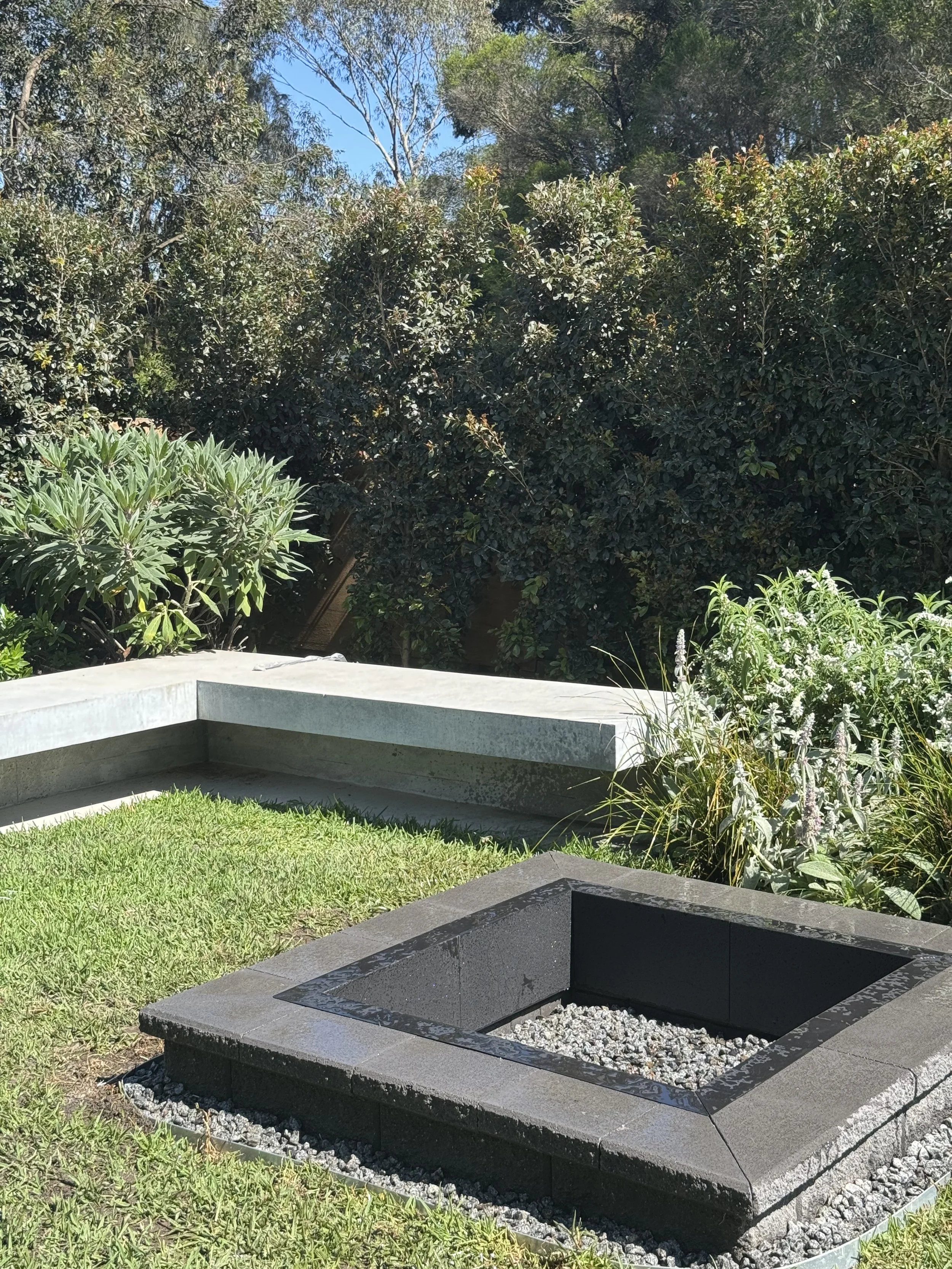 A modern outdoor fire pit with a square black inner basin filled with small gray stones, surrounded by a black stone border, set in a garden with green grass and plants, with lush bushes and trees in the background under a blue sky.