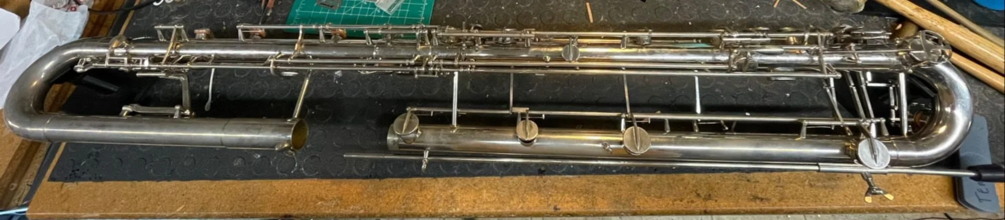 A disassembled silver leblanc paperclip contrabass clarinet lying on a black work surface with tools and workshop items nearby.