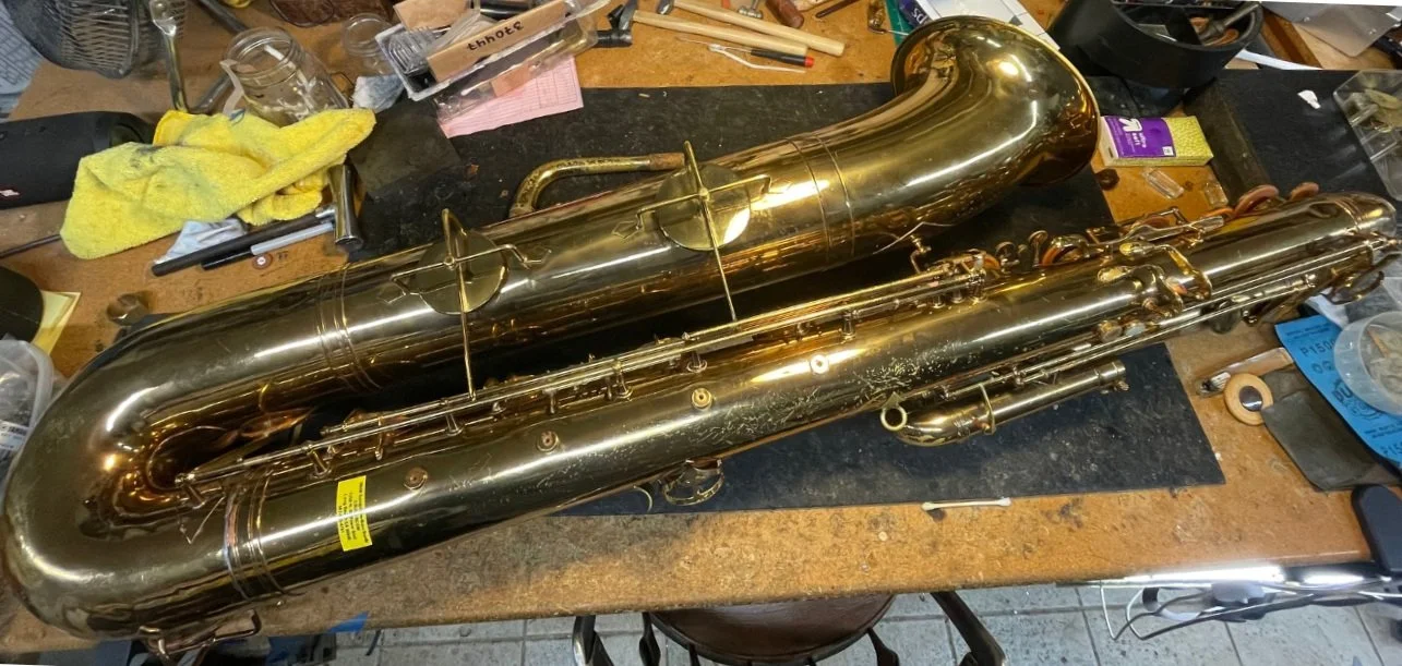 A bass saxophone lying on a cluttered workbench with tools and cleaning supplies around it.