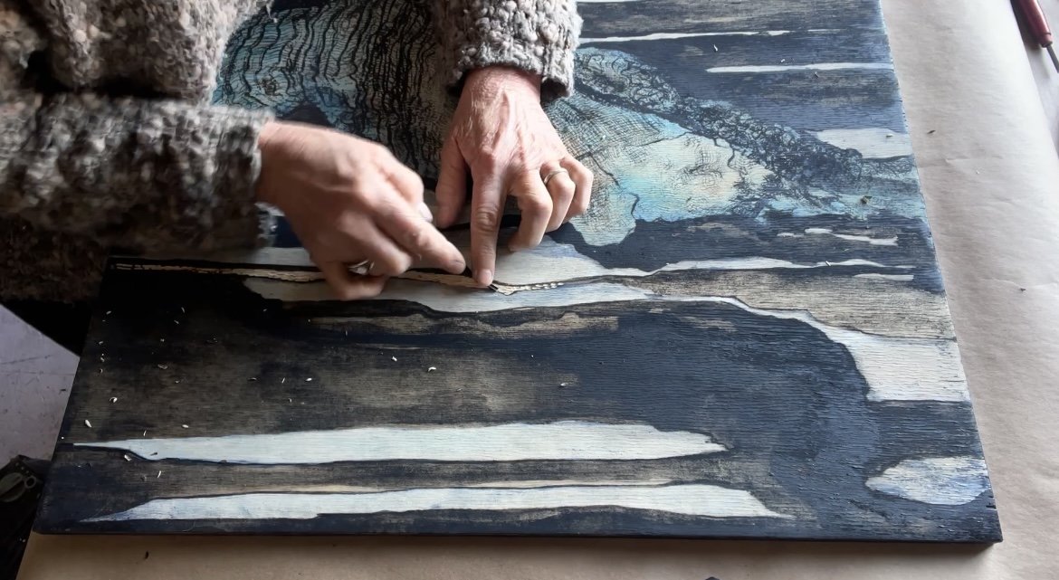 Person working on a wood art piece, using their hands to manipulate the wood surface with intricate detail. The artwork features natural wood textures with dark and light contrasting areas.
