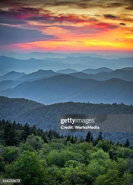 A scenic view of layered mountain ranges during sunset with colorful sky and lush green trees in the foreground.